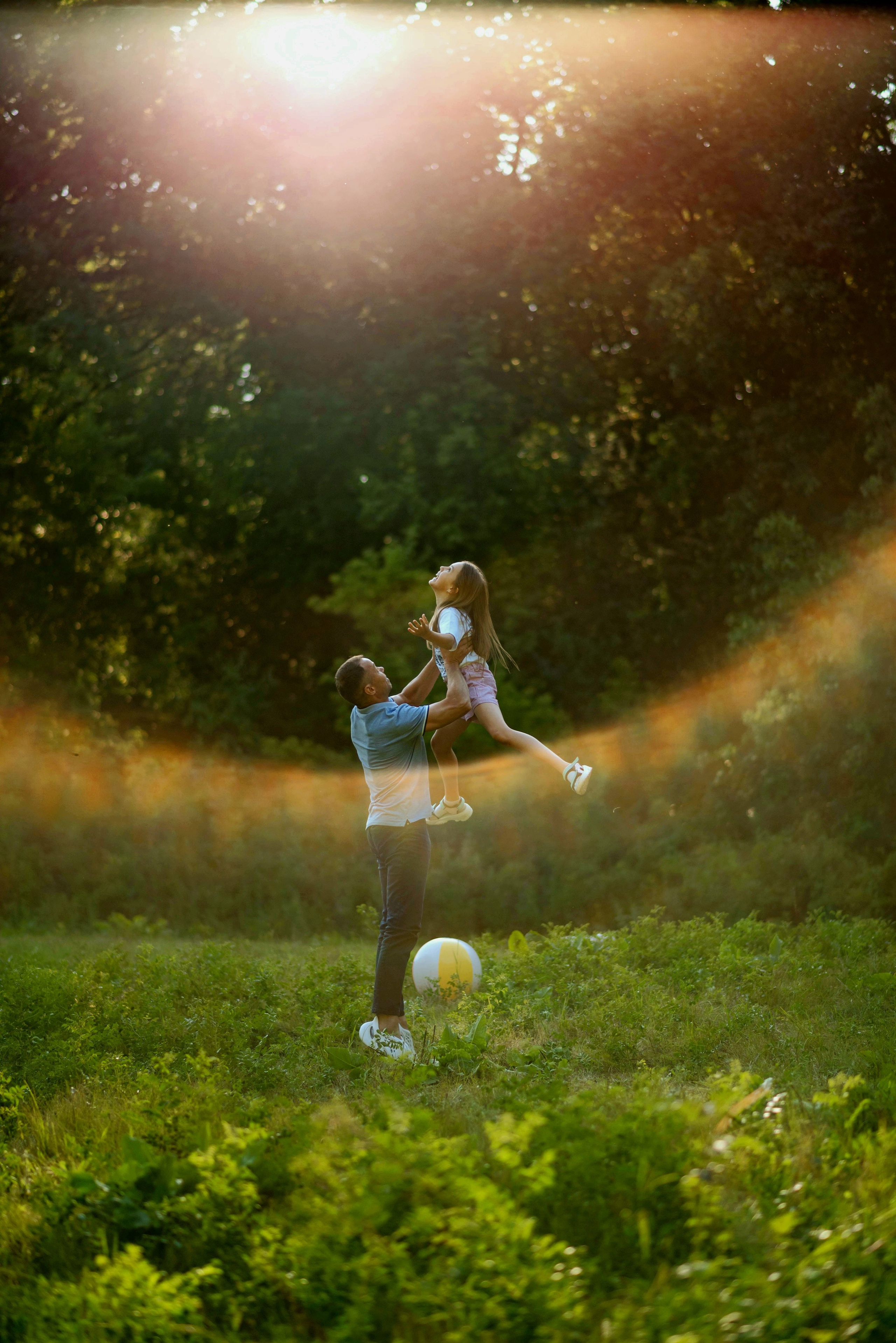 Отец держит дочку высоко над головой. На них падают солнечные лучи.