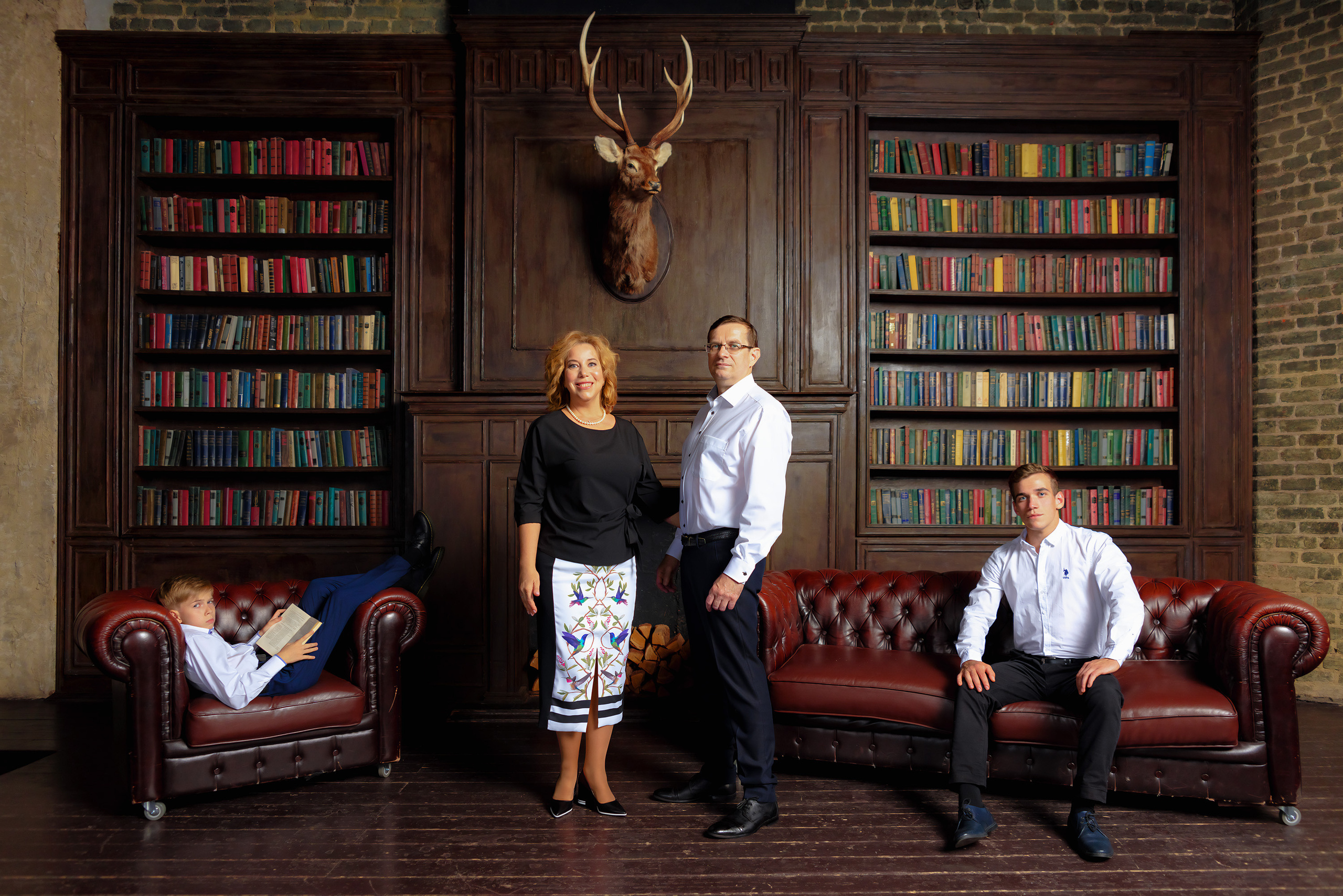 Родители стоят на фоне камина, книжных полок и головы оленя, сыновья сидят с двух сторон