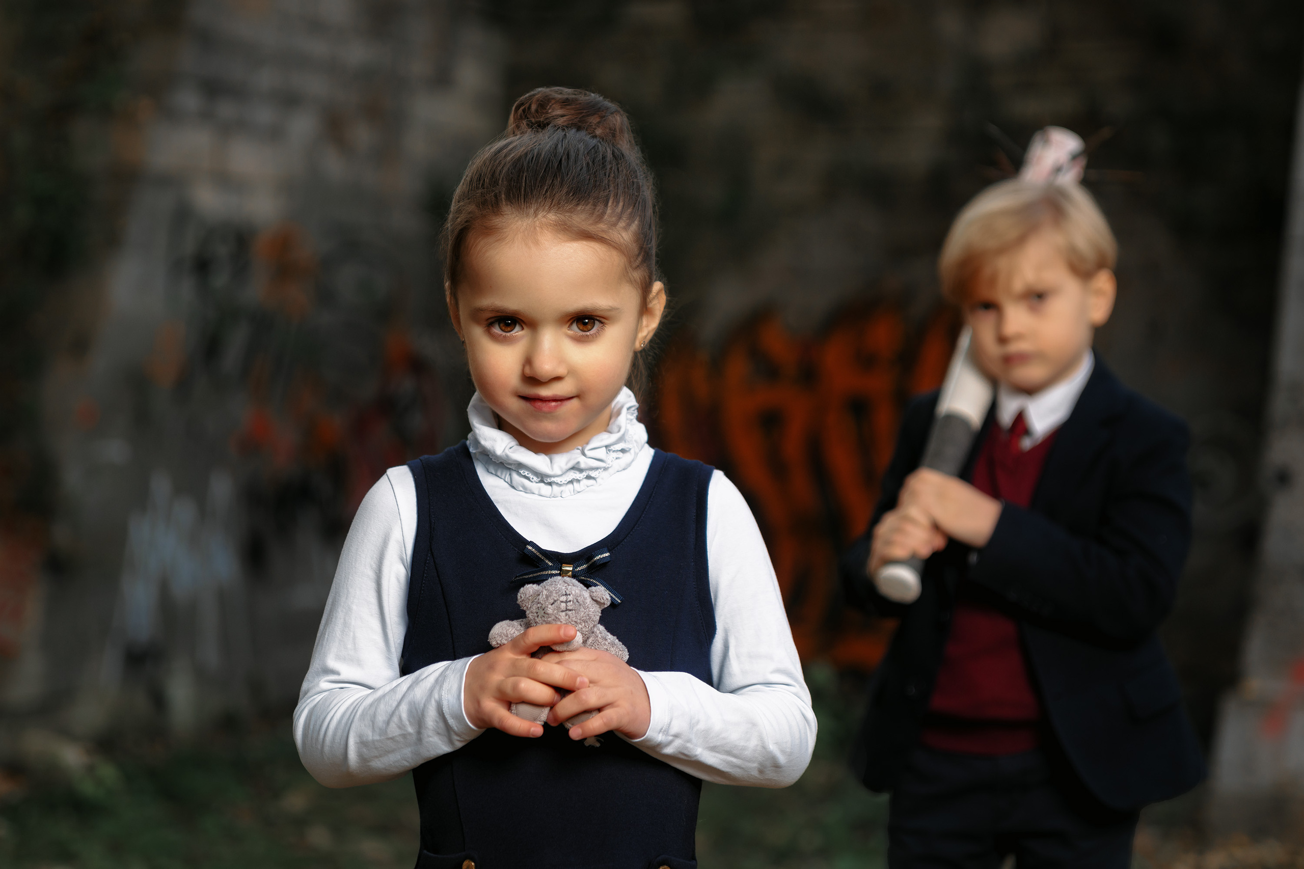 Девочка в школьной форме с мишкой в руках  смотрит в камеру на переднем плане, на заднем школьник с битой в руках