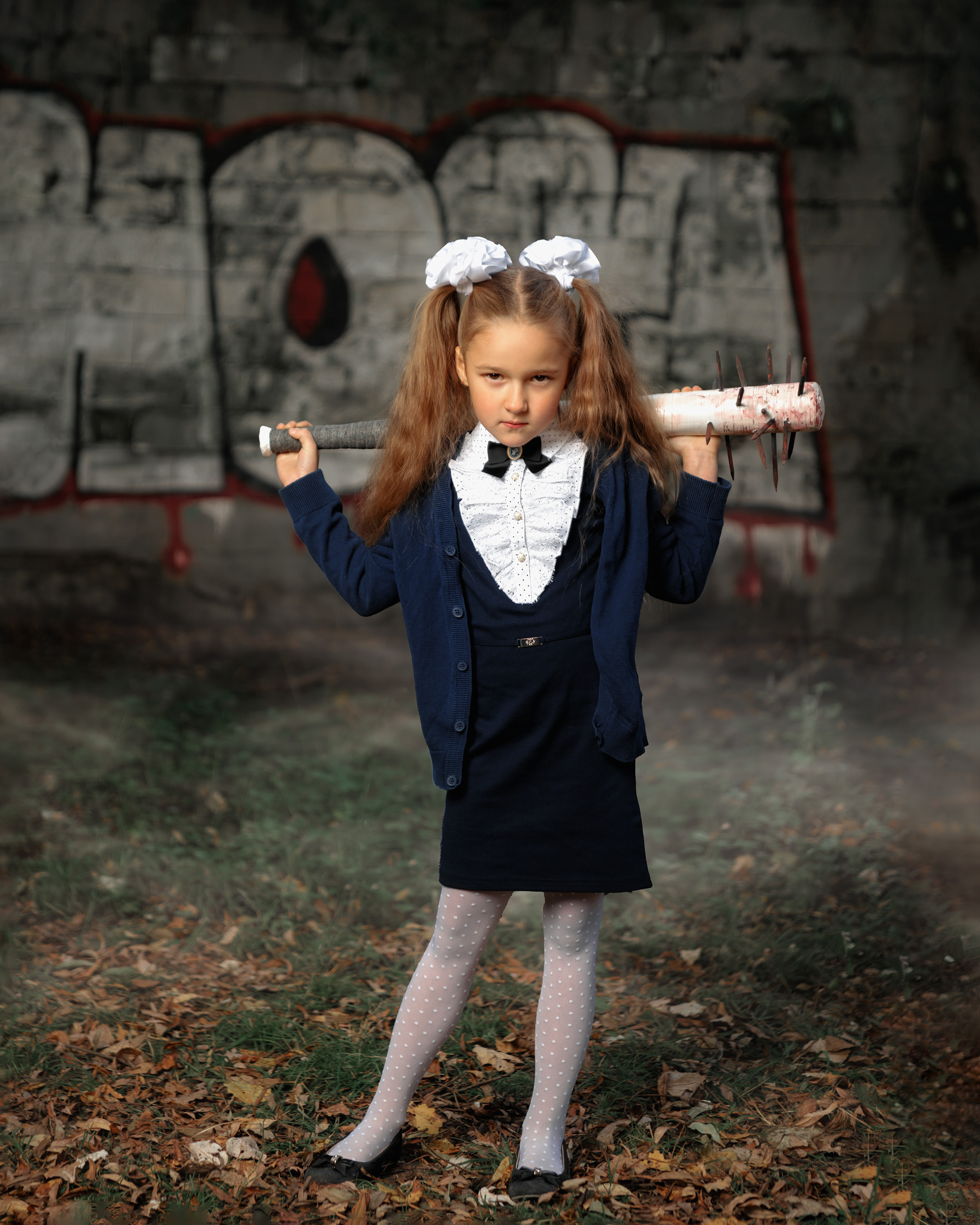 Девочка с белыми бантами в школьной форме с битой из зомби апокалипсиса на плечах на фоне стены с графити