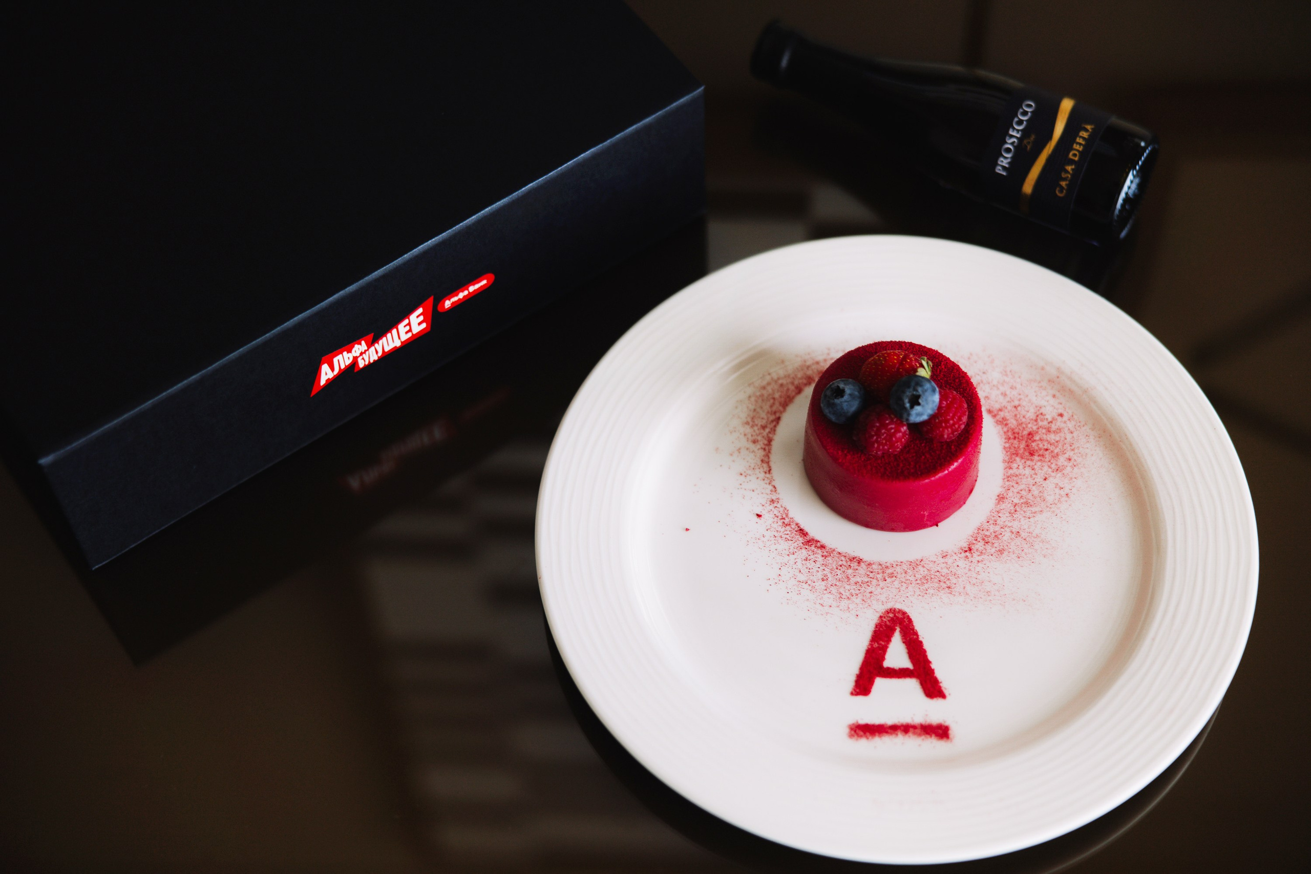 Детали: Фирменный десерт с логотипом «А» на белой тарелке рядом с брендированным подарком.