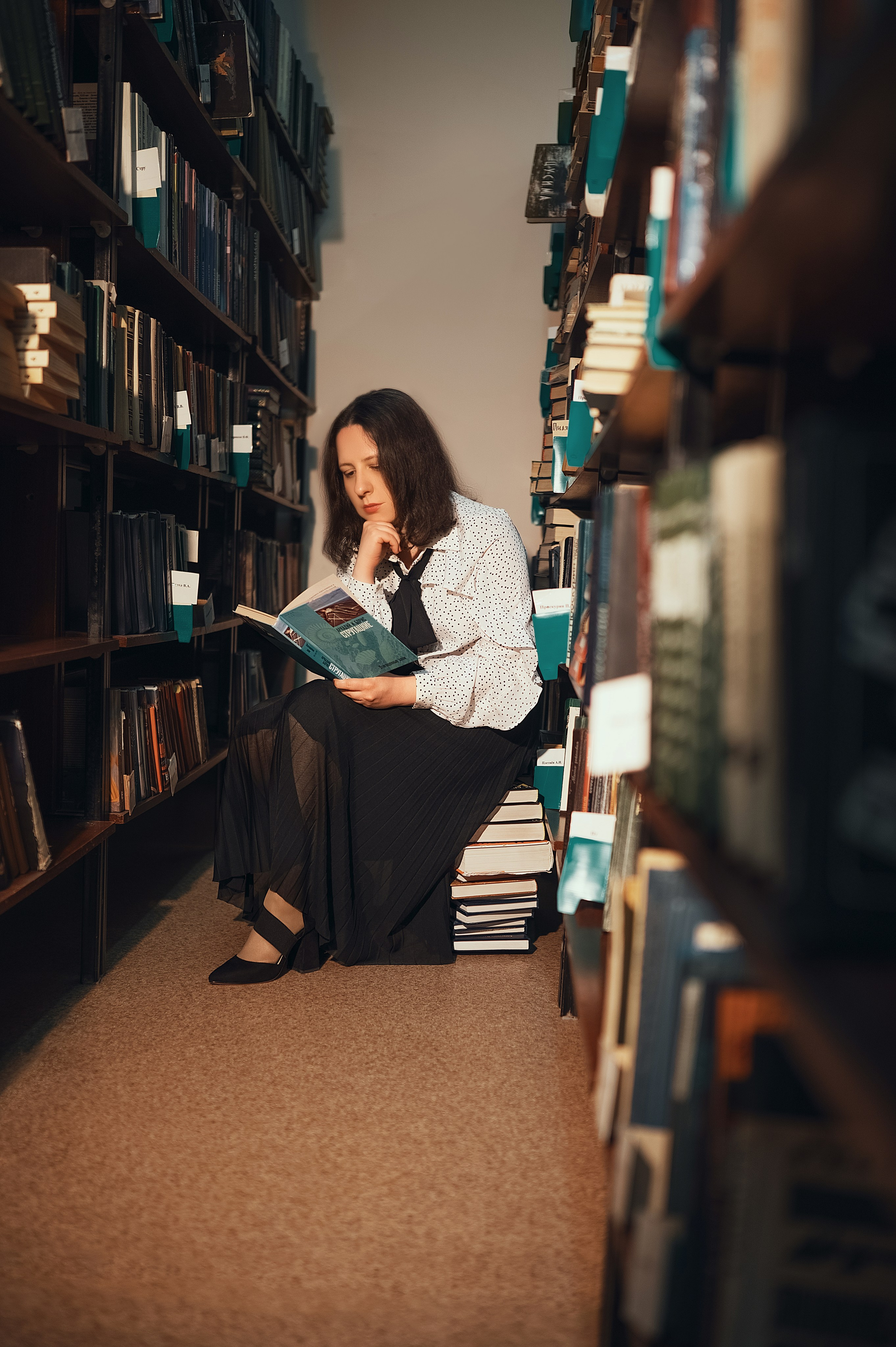 Фотосессия в Центральной библиотеке Волгодонска. Девушка в светлой блузке и тёмной юбке читает книгу среди стеллажей. Фотограф — Елизавета Жуйкова.