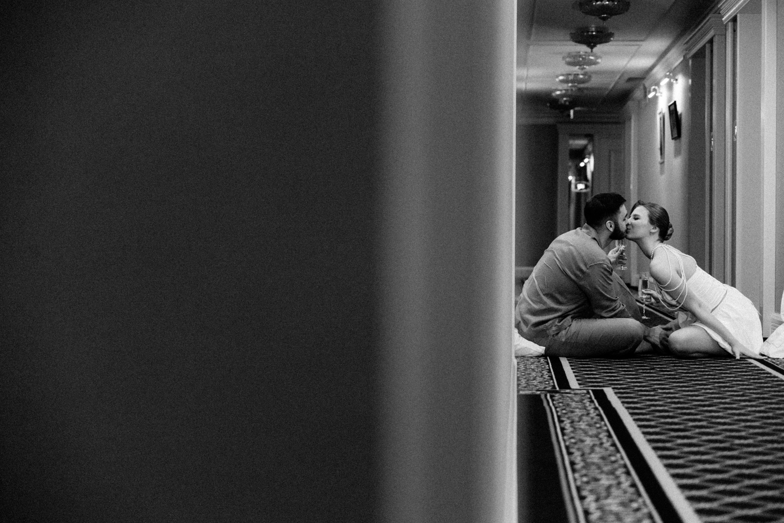 Черно-белое фото жениха и невесты, сидящих на полу в коридоре.