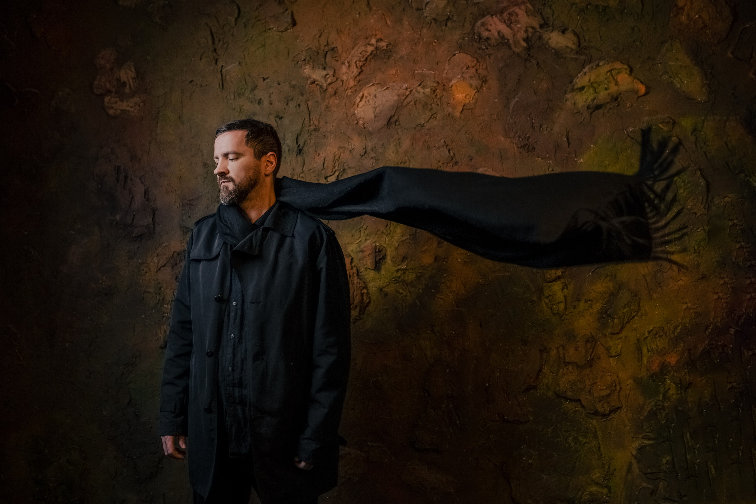 Портрет брутального мужчины с бородой и в черном пальто с развивающимся шарфом.