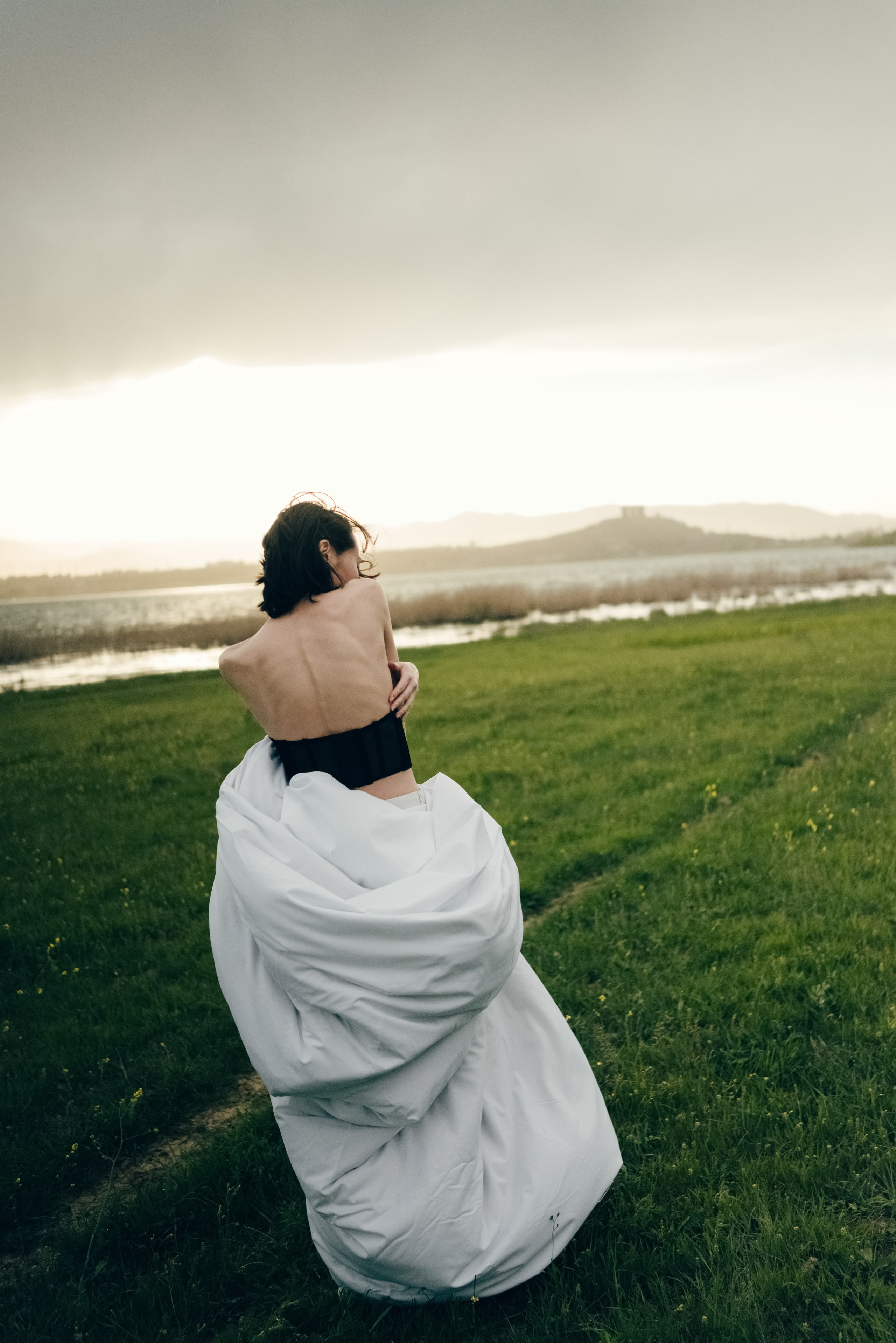 Фото девушки стоящей с нам спиной на фоне заката над водой. Девушка стоит в поле и закутана в теплое белое одеяло