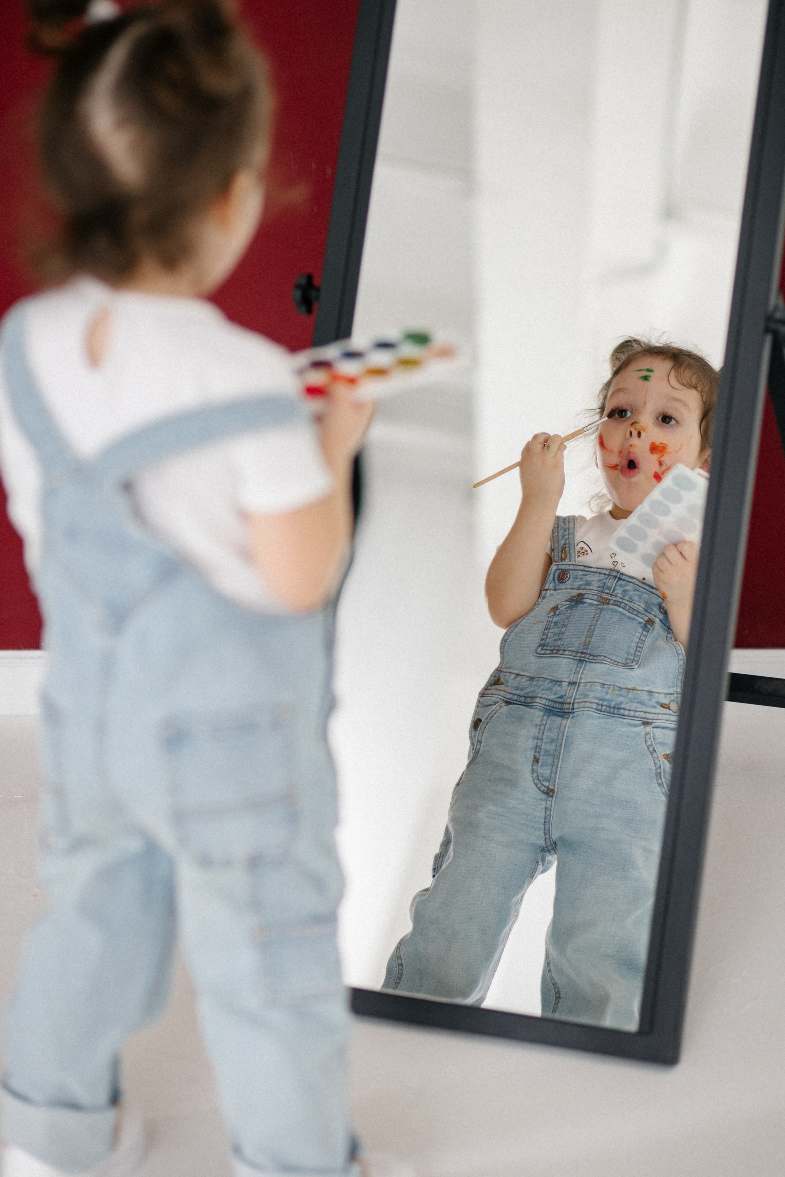 Краски на лице и холсте: девочка дурачится, раскрашивая свое лицо перед зеркалом. Студийная съёмка с юмором и теплом