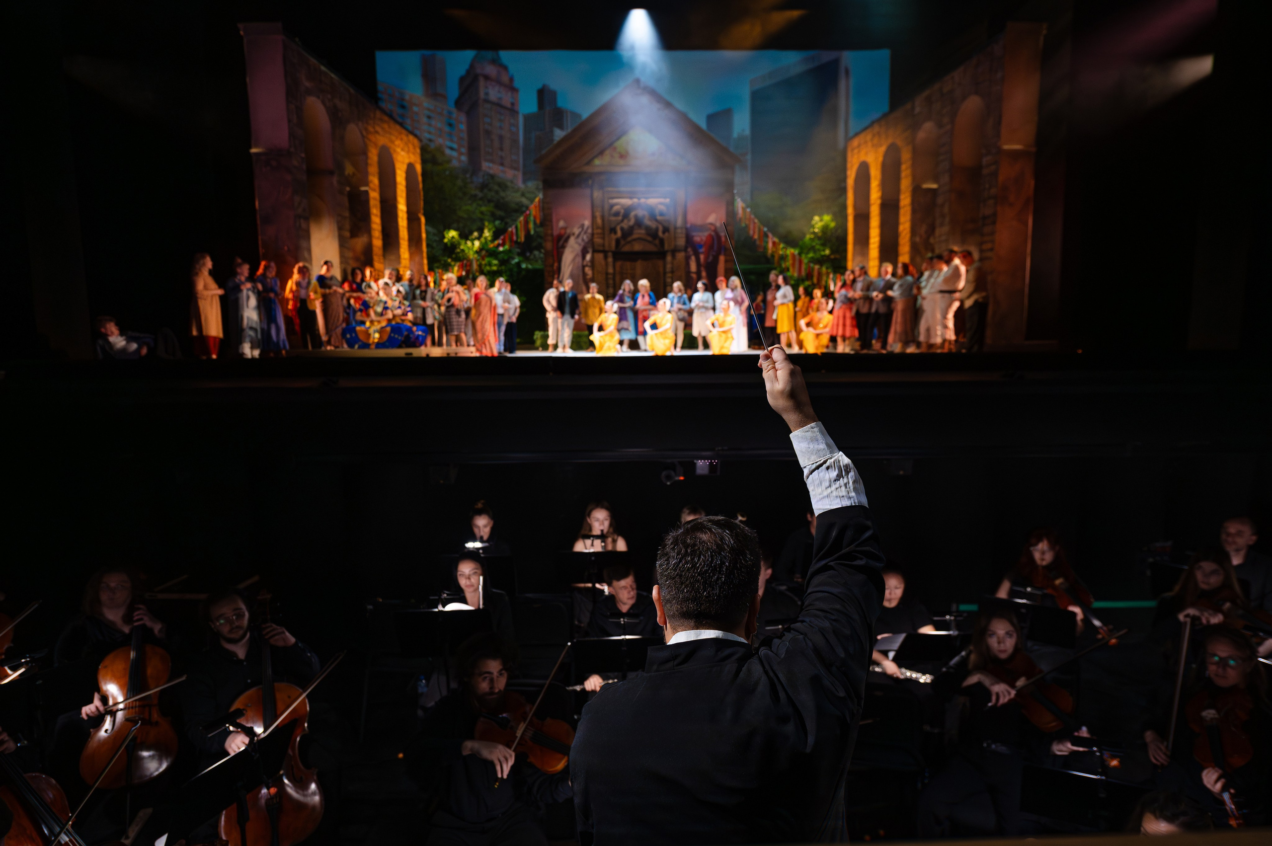 Дирижёр в инструментальной яме с оркестром поднимает палочку на фоне идущего представления оперы на сцене 
