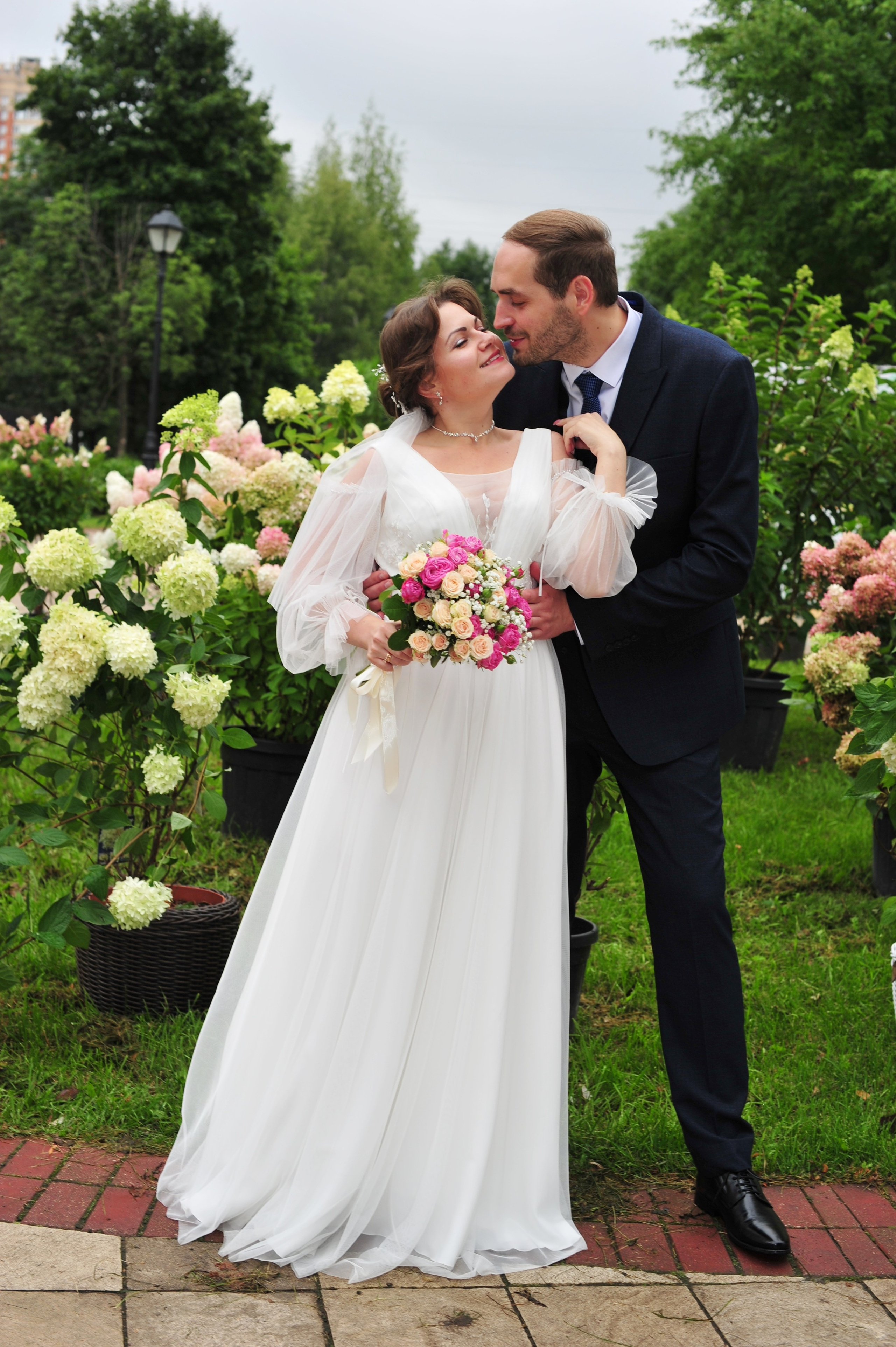 Свадьба в Царицыно пасмурным летним днем, портрет новобрачных в полный рост на фоне цветущих гортензий в парке 