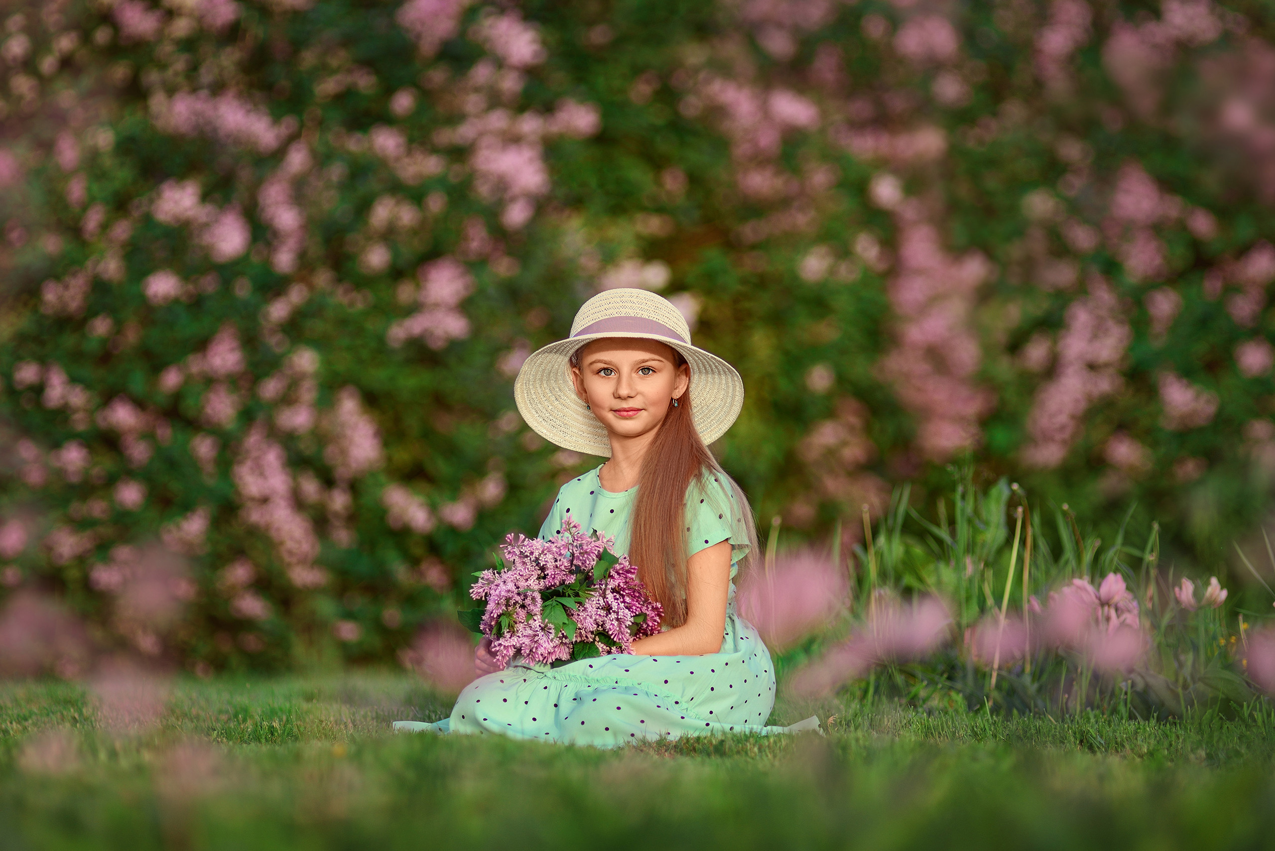 Девочка в широкополой шляпе сидит на траве с букетом сирени на фоне сиреневого куста.