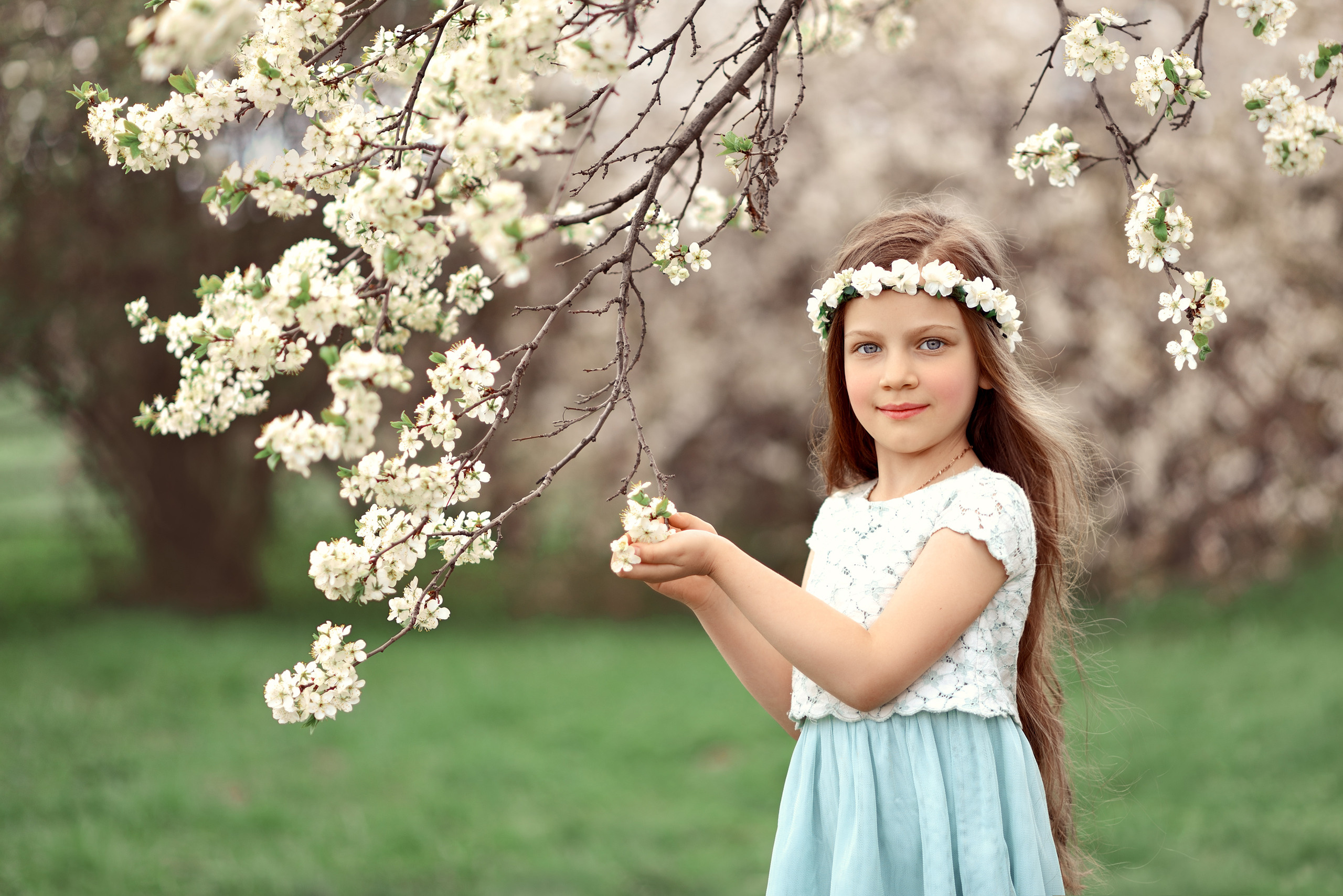 Девочка в белом венке трогает ветви цветущей яблони.