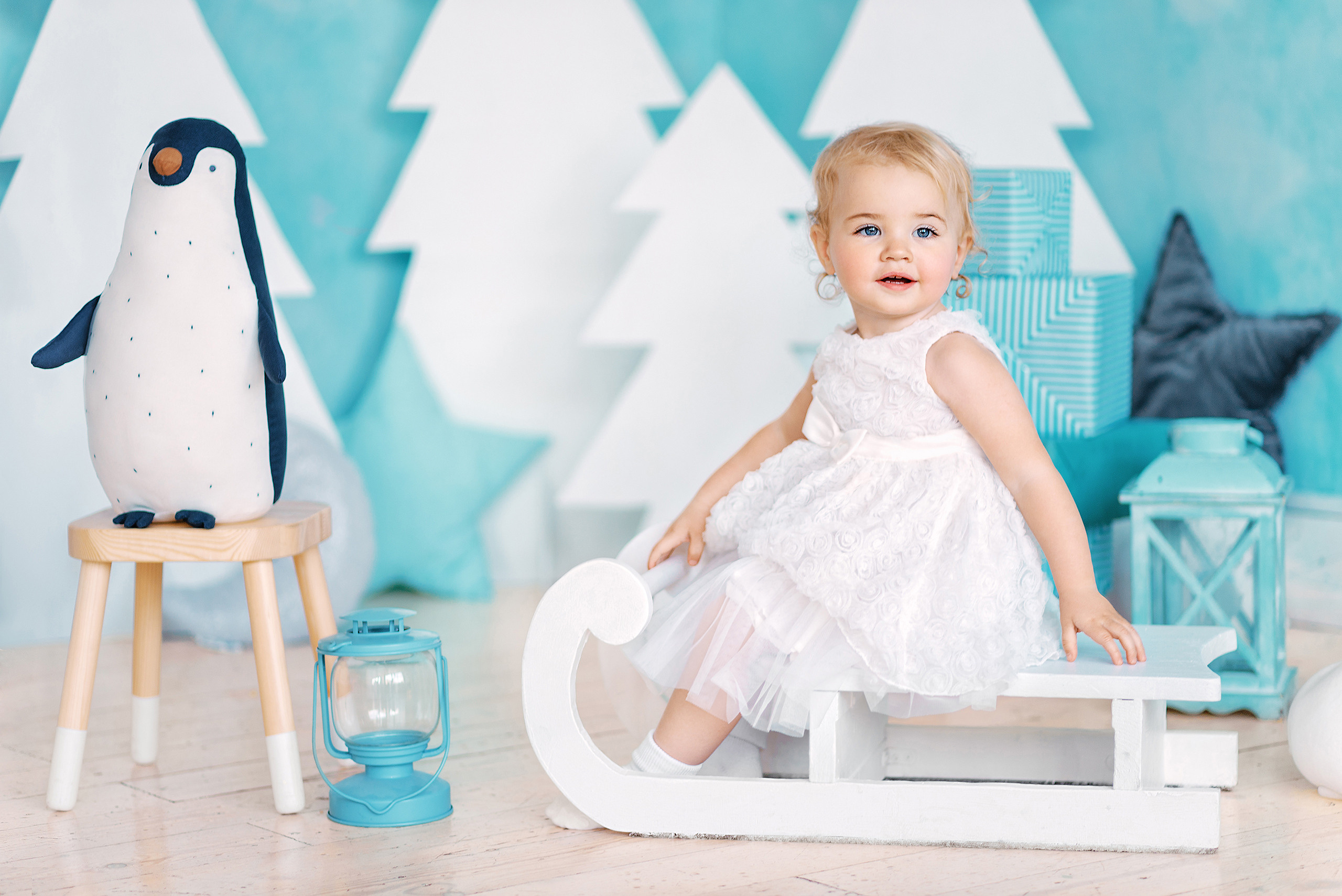 Девочка в новогодней фотостудии сидит на деревянных санках рядом с игрушечным пингвином.