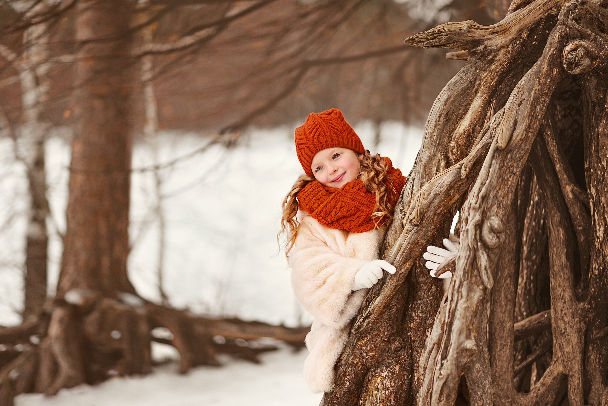 Девочка в белой шубке выглядывает из-за дерева во время зимней прогулки.