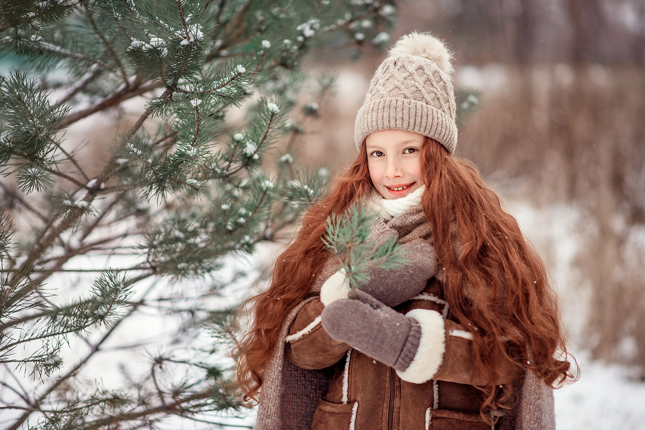 Девочка с длинными рыжими волосами держит еловую веточку на фоне зимнего пейзажа.