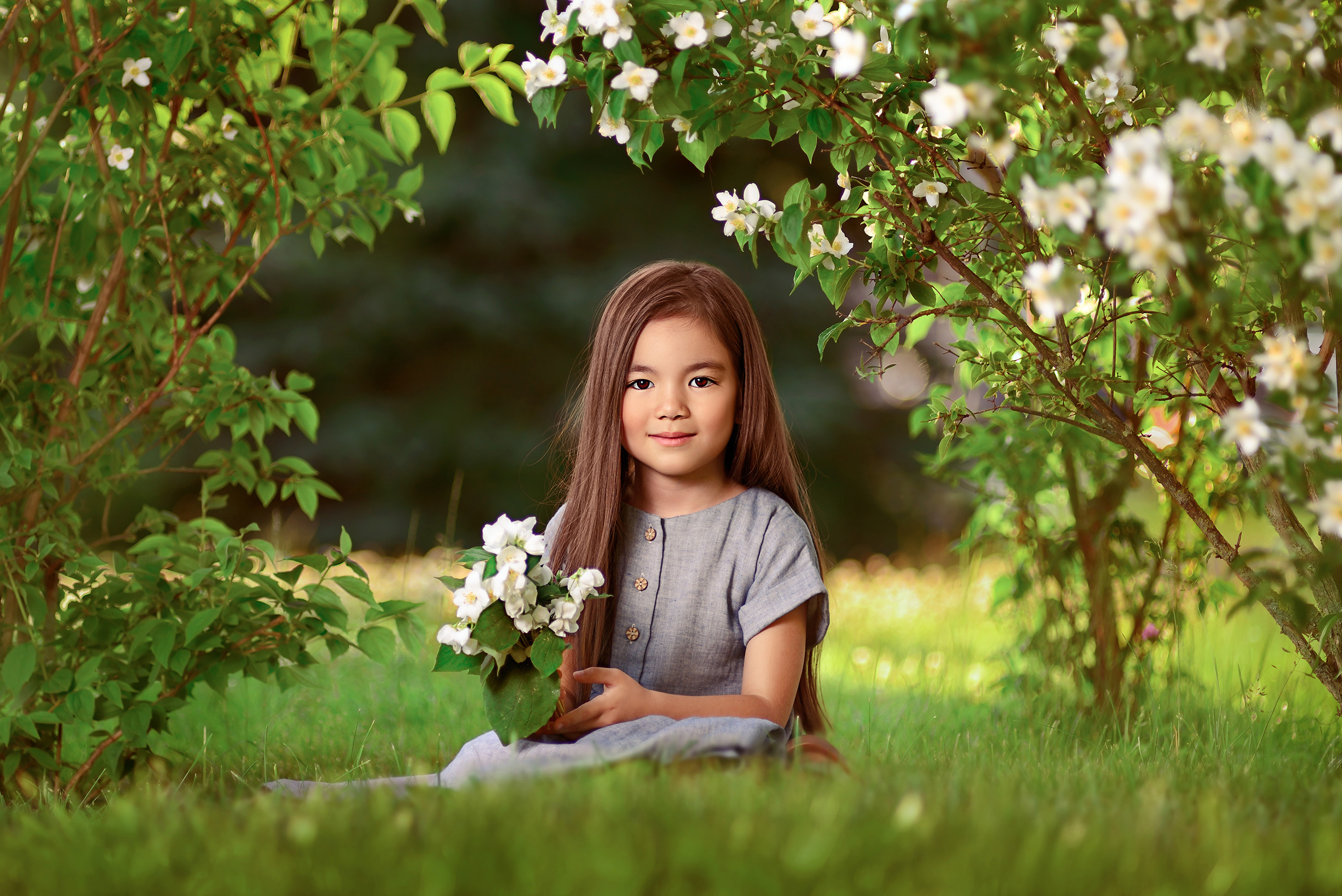 Девочка в кустах белой акации сидит на траве с букетом.