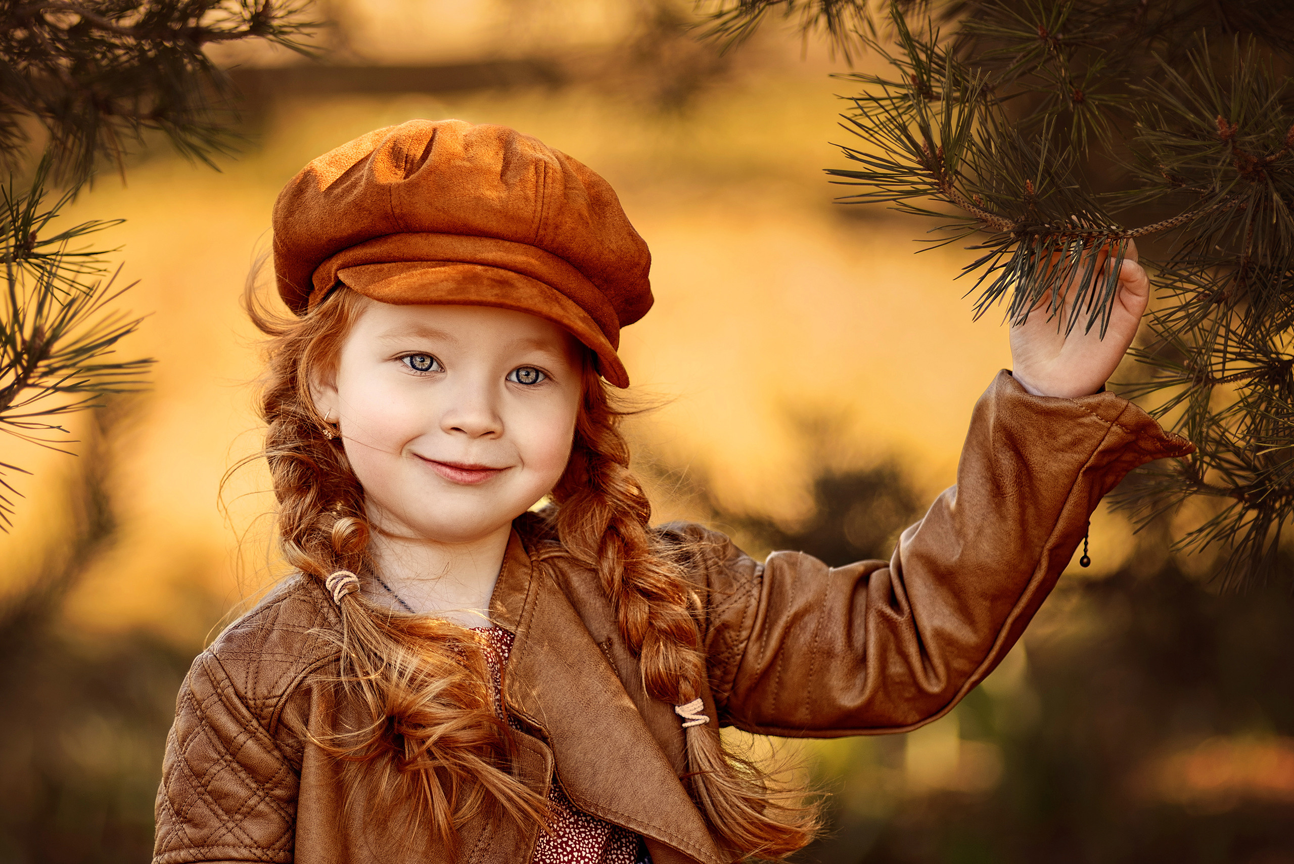 Рыжая девочка в кепке с двумя косами держится за еловую ветку.