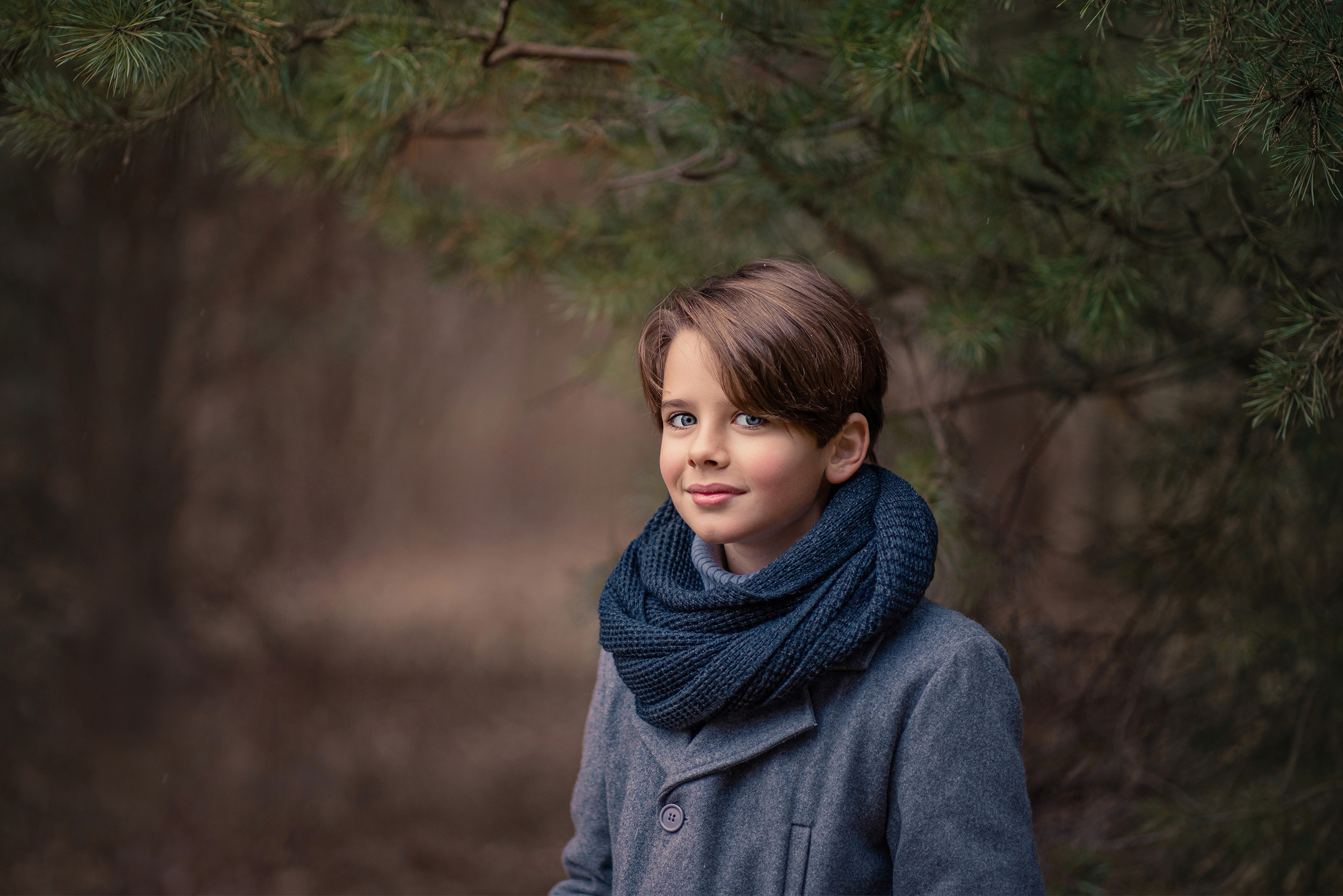 Портрет мальчика в сером пальто и шарфе на фоне елового леса.