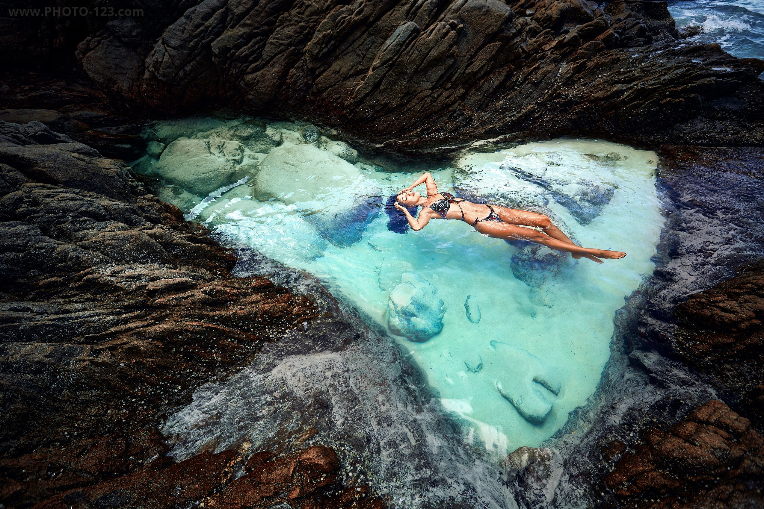 Woman in bikini floating on her back in a natural turquoise rock pool by the ocean, viewed from above, rugged dark rocks surrounding clear water, serene mood, tropical coastal landscape, luxury travel and nature escape concept