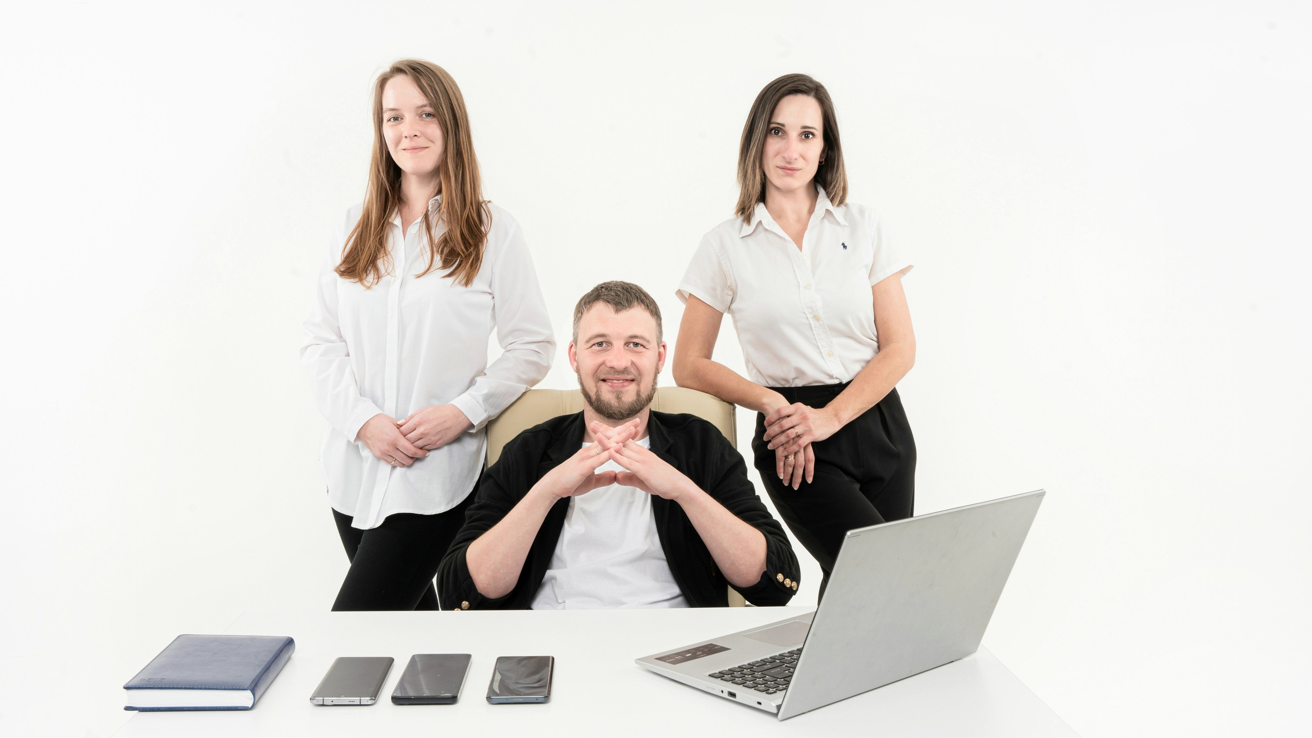 На фотографии изображено трое людей, сидящих за белым столом. Перед центральной фигурой открыт ноутбук, а слева расположены три смартфона и, возможно, блокнот или планер, аккуратно выложенные в ряд. Фон белый, все люди одеты в деловой кэжуал. Человек посередине скрещивает пальцы в стиле "венец" (как у Мистера Бернса из "Симпсонов"), а остальные держат руки по-разному: один на колене, а другой на столе.