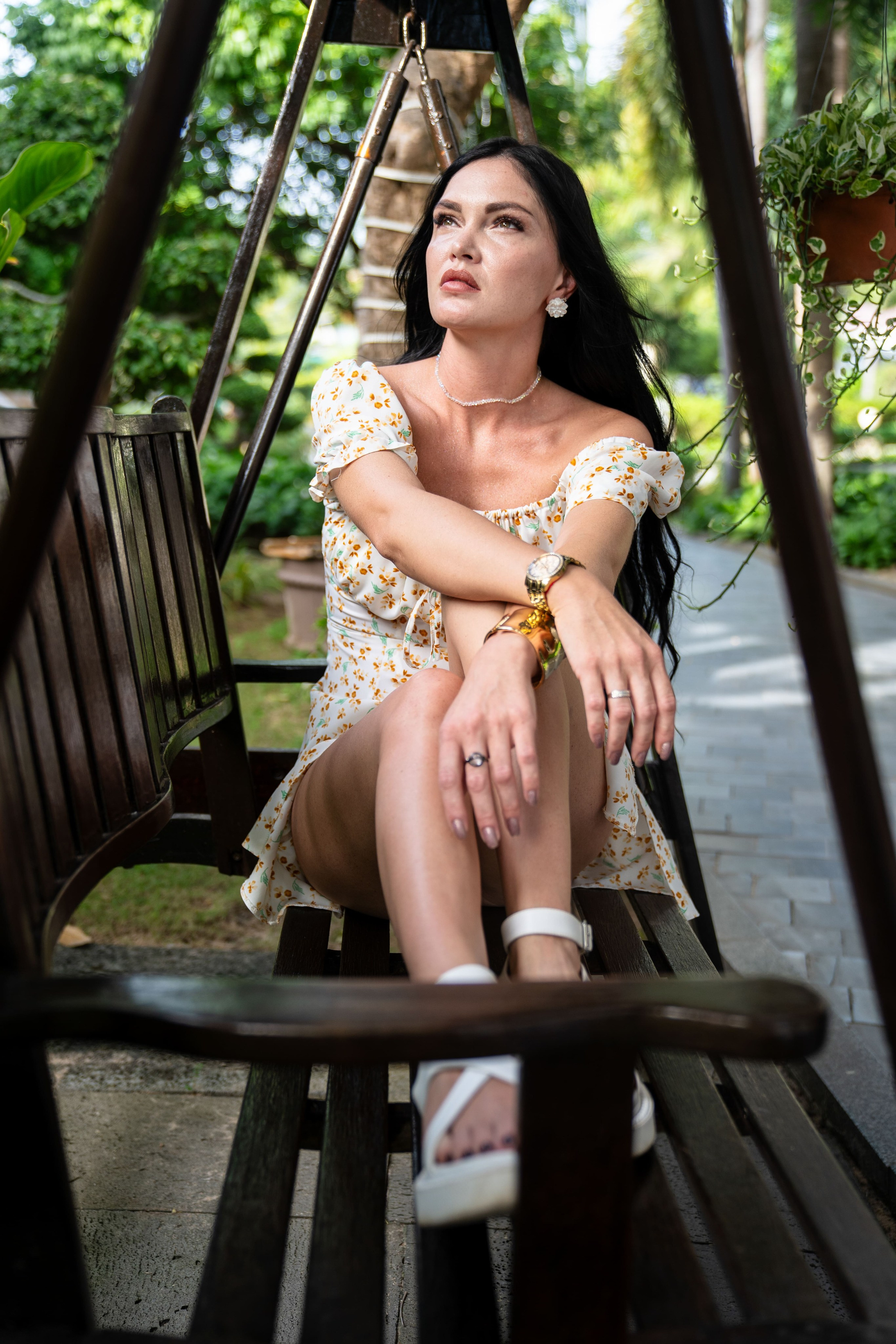 Женский портрет в летнем платье: девушка сидит на массивных деревянных качелях в саду.