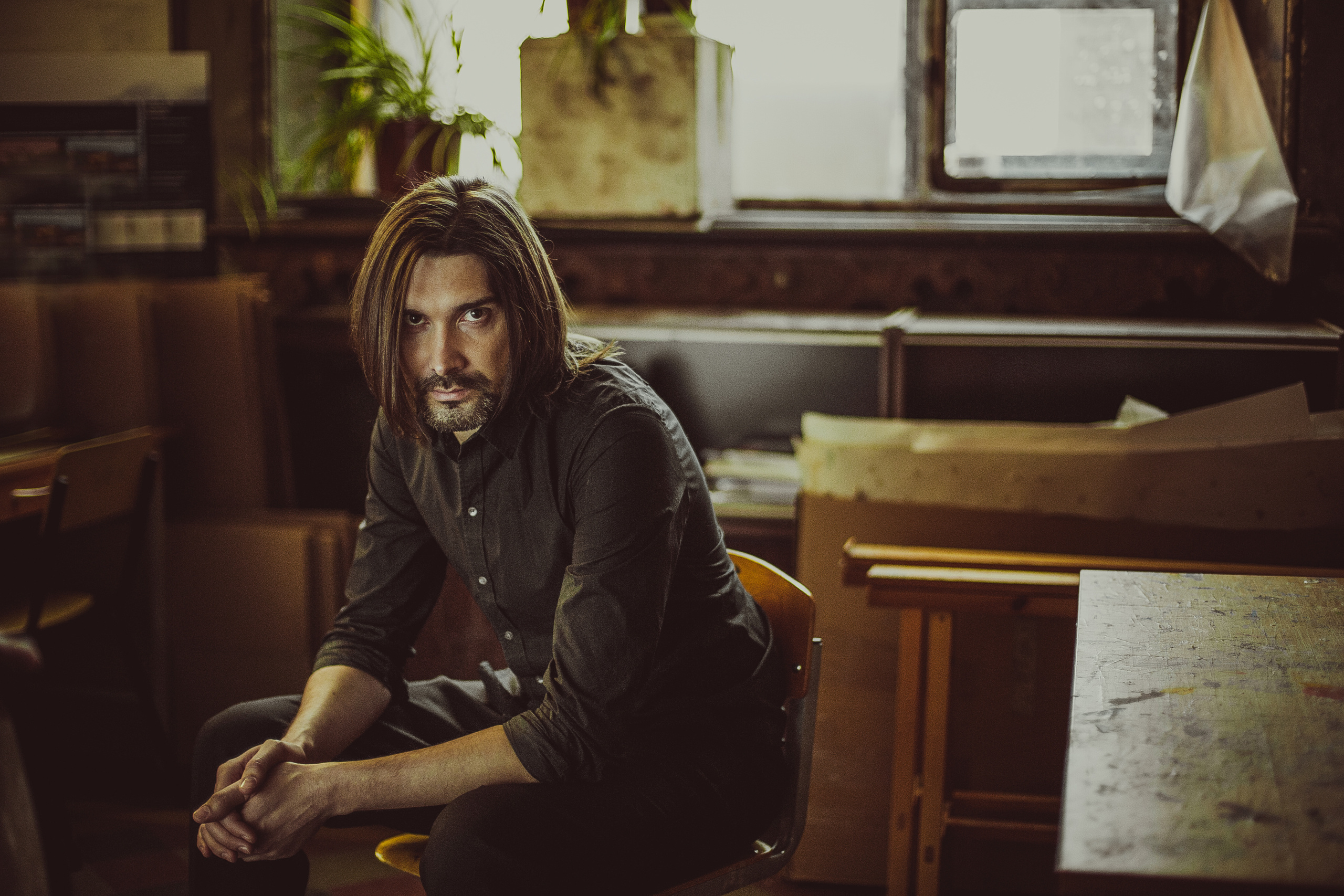 портрет брутального длинноволосого мужчины в темной рубашке сидящего на стуле
