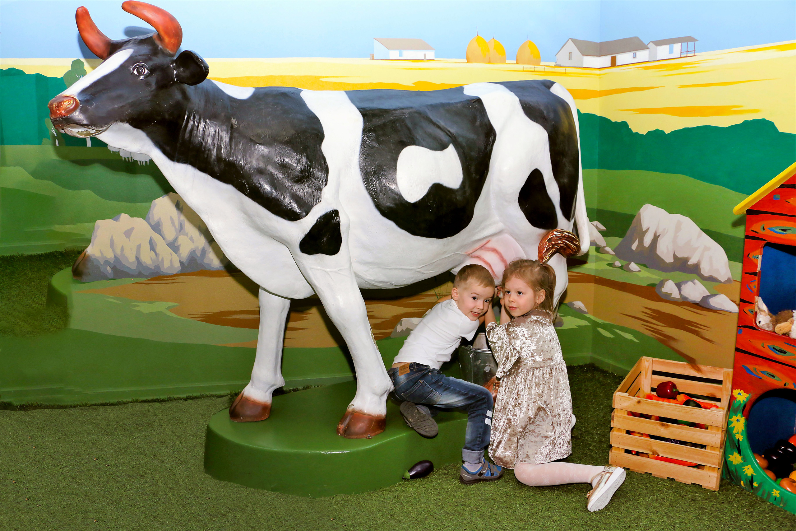 Корова стоит. Рядом с коровой мальчик и девочка. Девочка доит корову, а мальчик помогает девочки