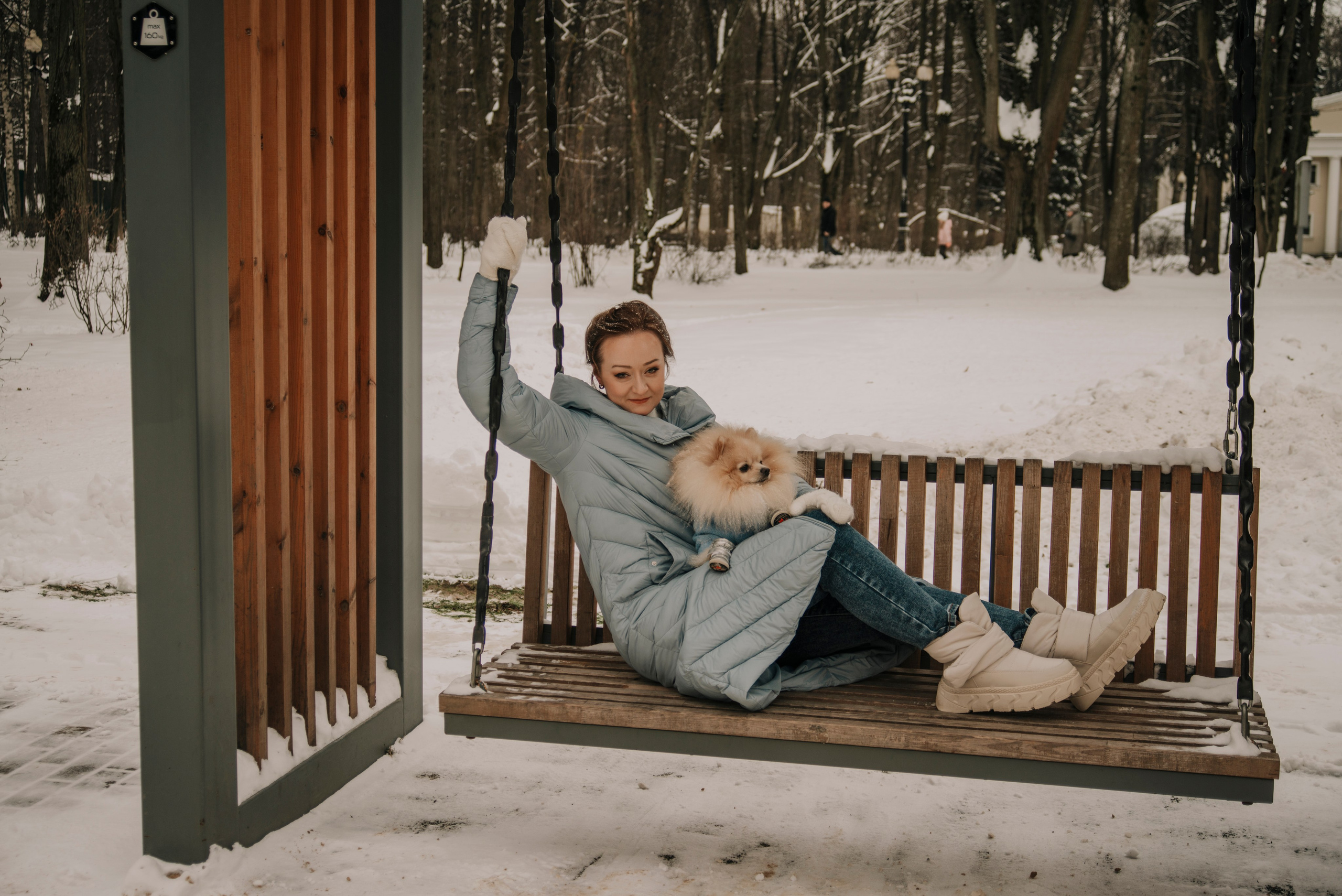 Девушка в голубом пуховике сидит на качелях в зимнем парке и держит на руках собаку шпица