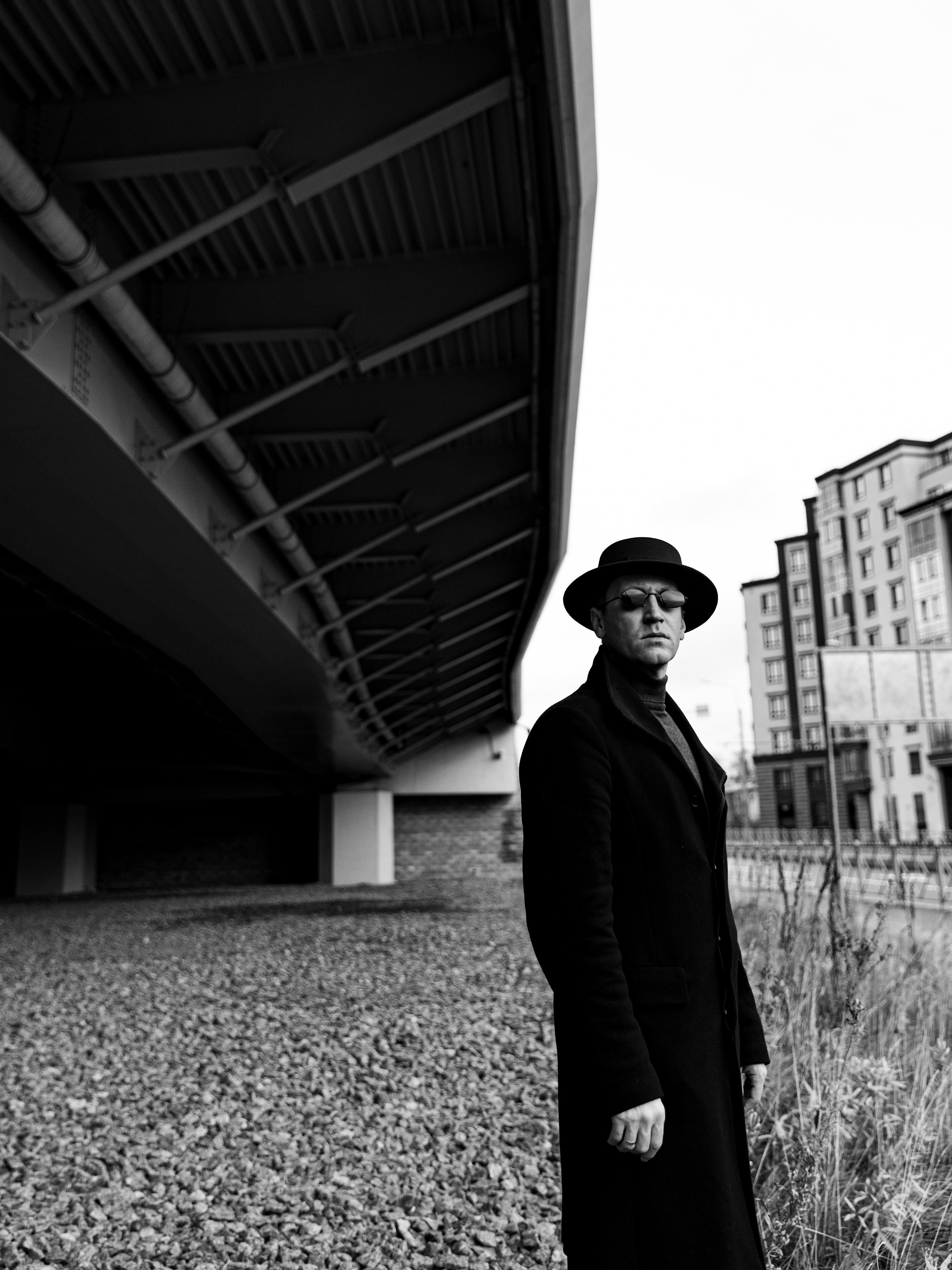 Черно-белый портрет мужчины в пальто, шляпе и очках у моста