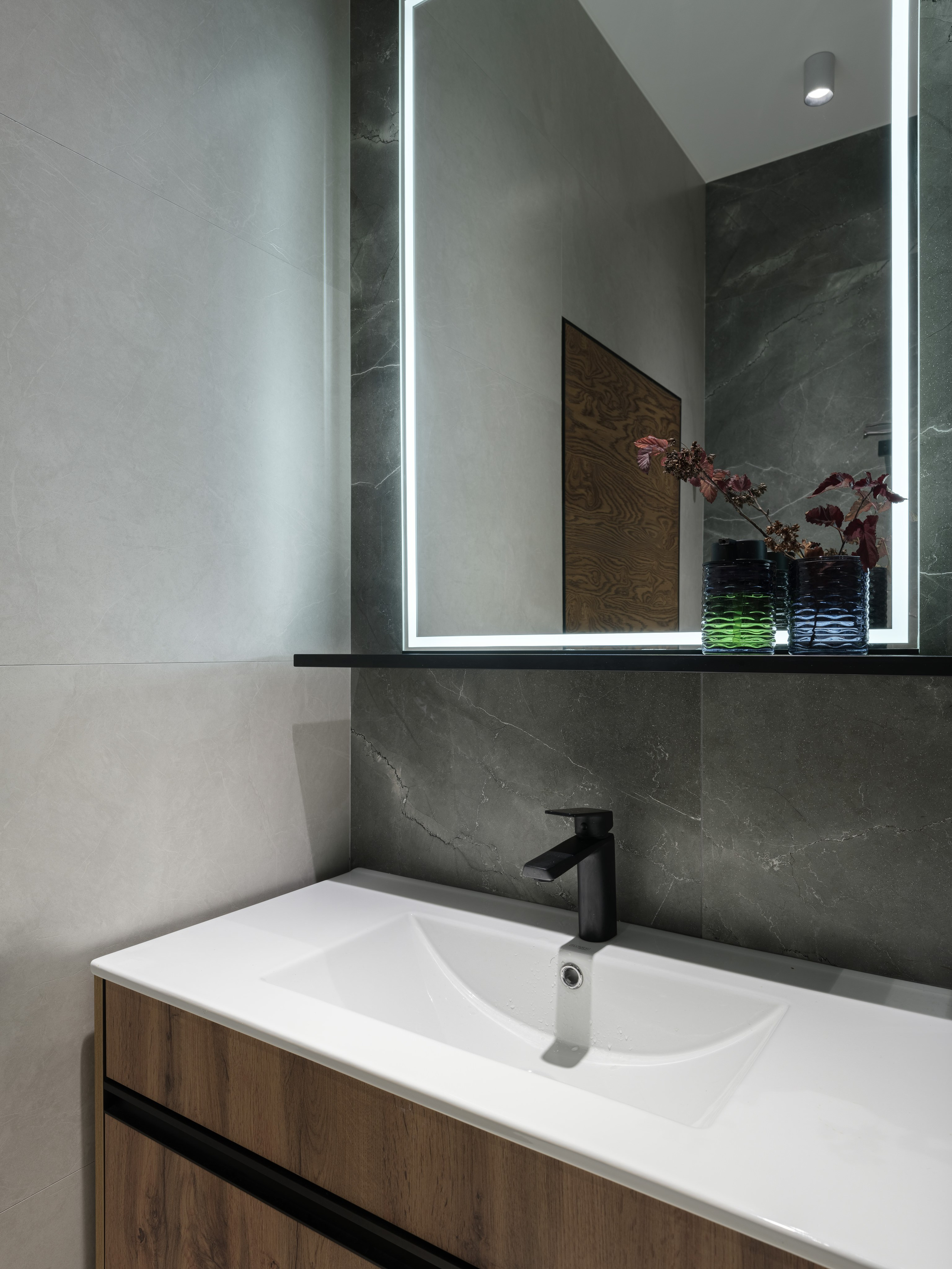 Фрагмент современной ванной комнаты в стиле минимализм с подсвеченным зеркалом, белой раковиной и тумбой из дерева, черным смесителем и акцентом на серую плитку под камень. Интерьер оформлен дизайнером Алиной Дёровой, стилист Наталья Якушева, фото Сергей Денисенко.