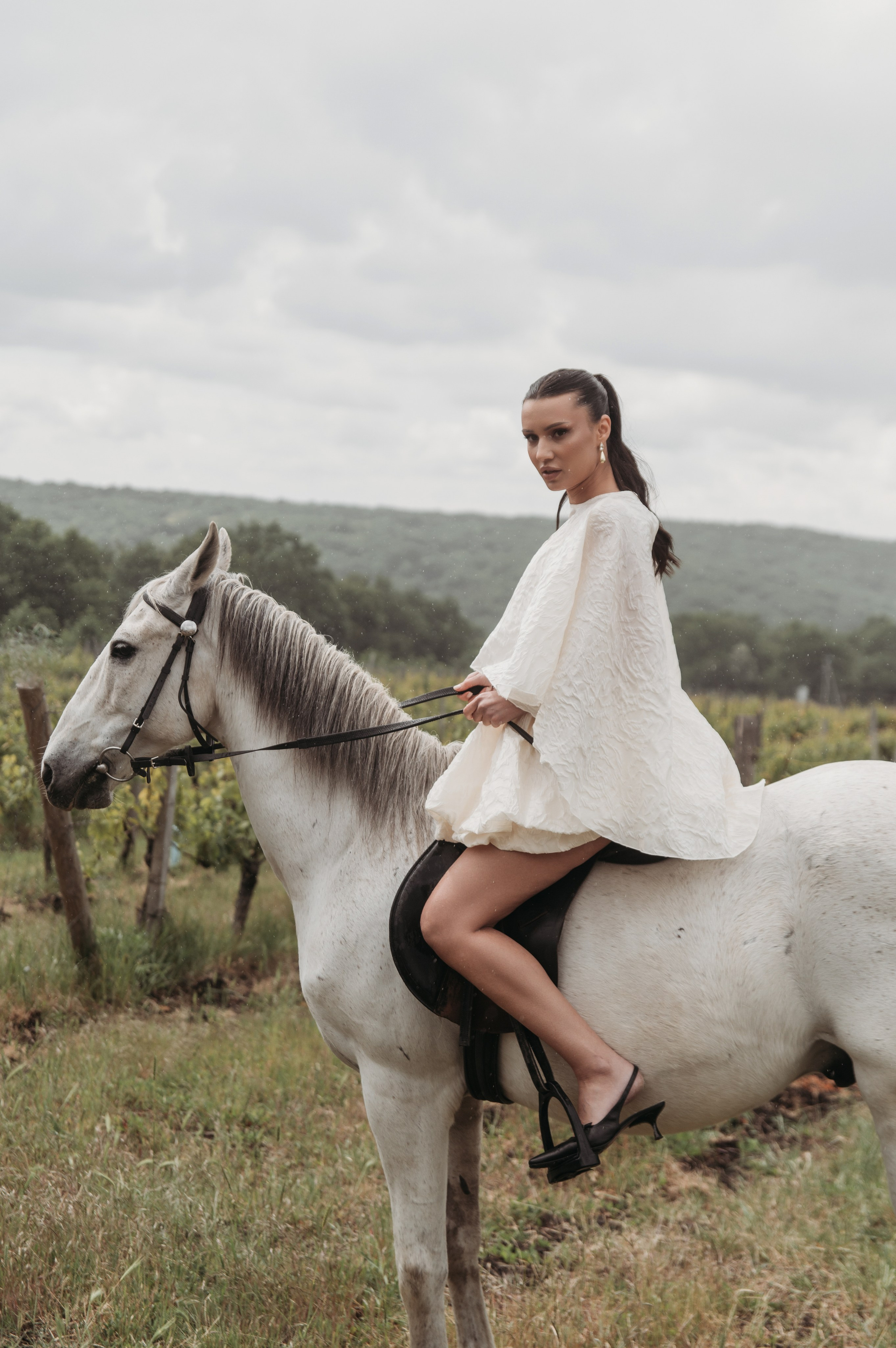 Невеста в белом свадебном платье сидит верхом на белой лошади, с легкой улыбкой и романтичным образом, на фоне виноградника и облачного неба