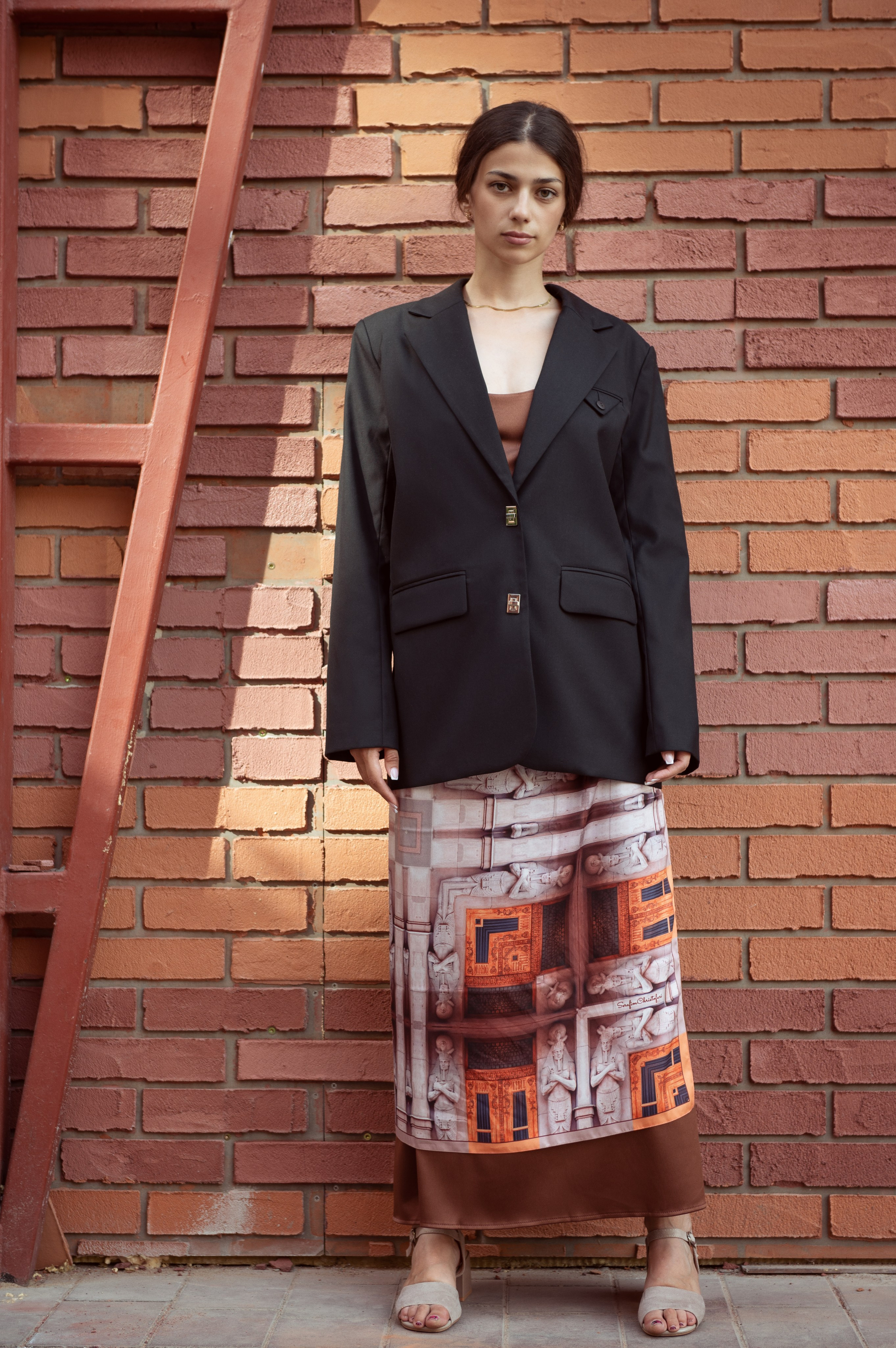 Красивая темноволосая девушка в черном пиджаке стоит на фоне кирпичной стены, на ней шелковый платок от Serafim Cristofari "Египетский дом". Пространство Seno
