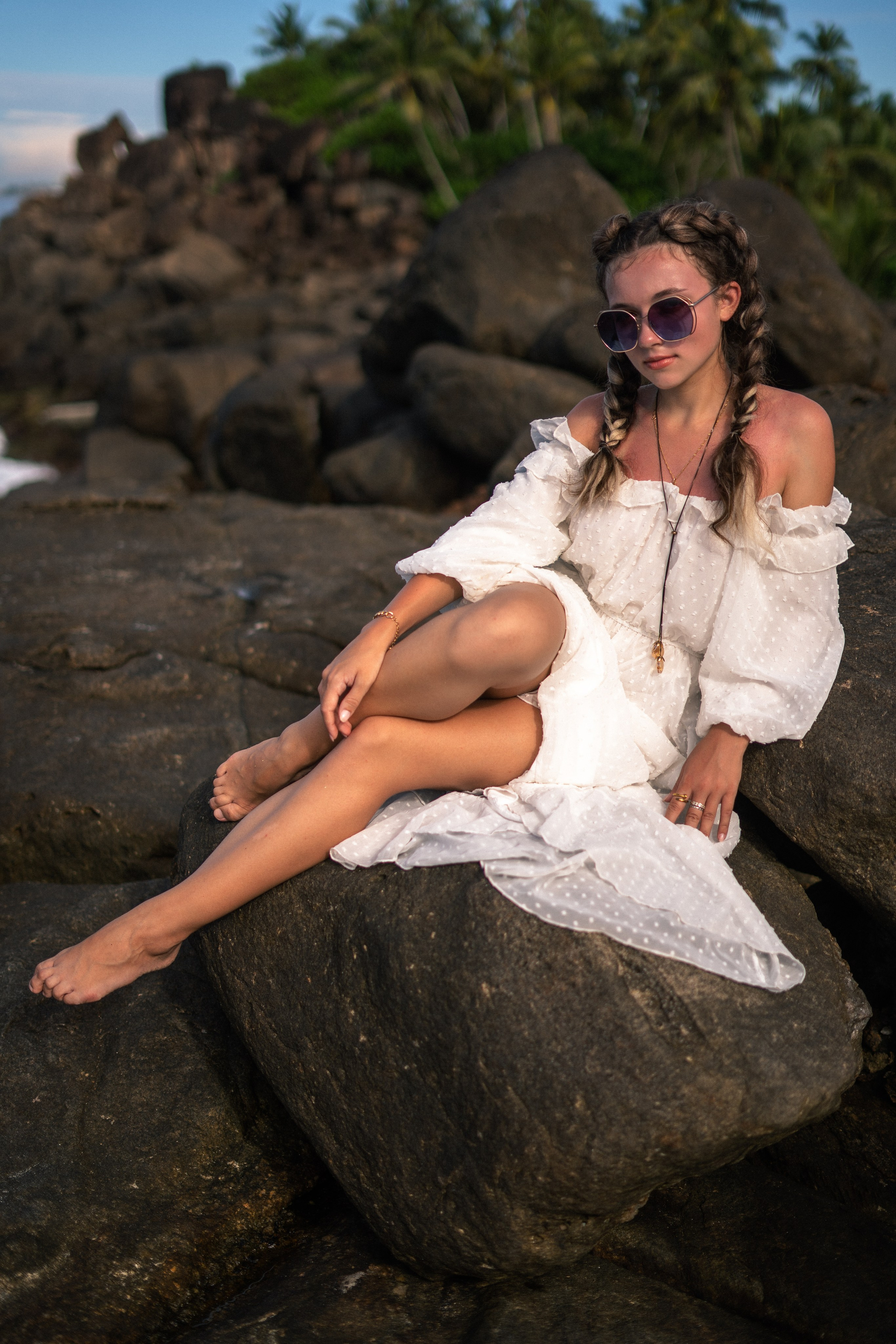 a girl in a white dress and glasses sitting on the rocks with an ocean view