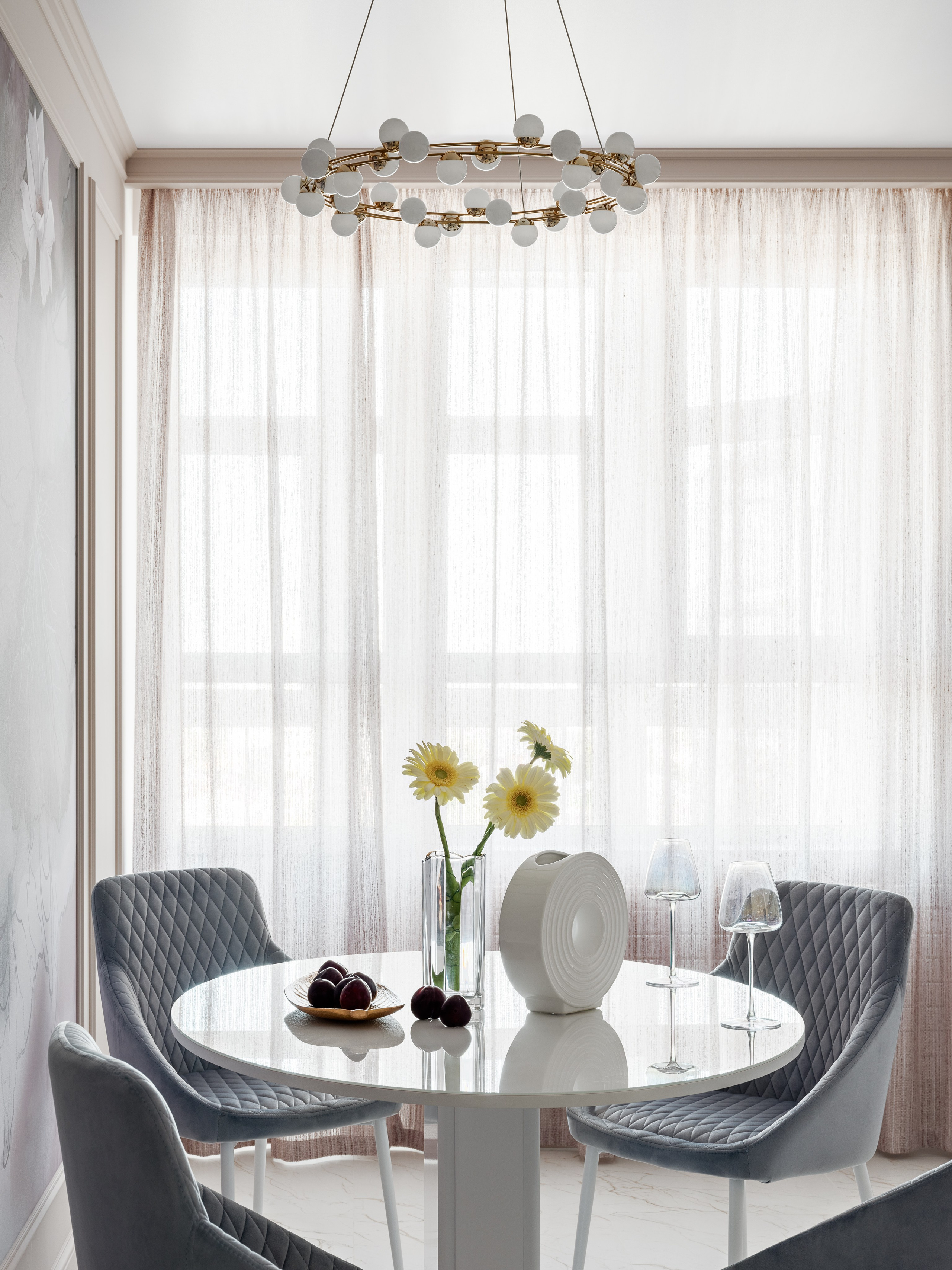 Уютная обеденная зона у окна с белым круглым столом, мягкими стульями серого цвета и стильным освещением, дополненная современным декором и цветами; атмосферный интерьер от дизайнера Натальи Бунеевой, стилиста Натальи Якушевой, фотографа Сергея Денисенко.