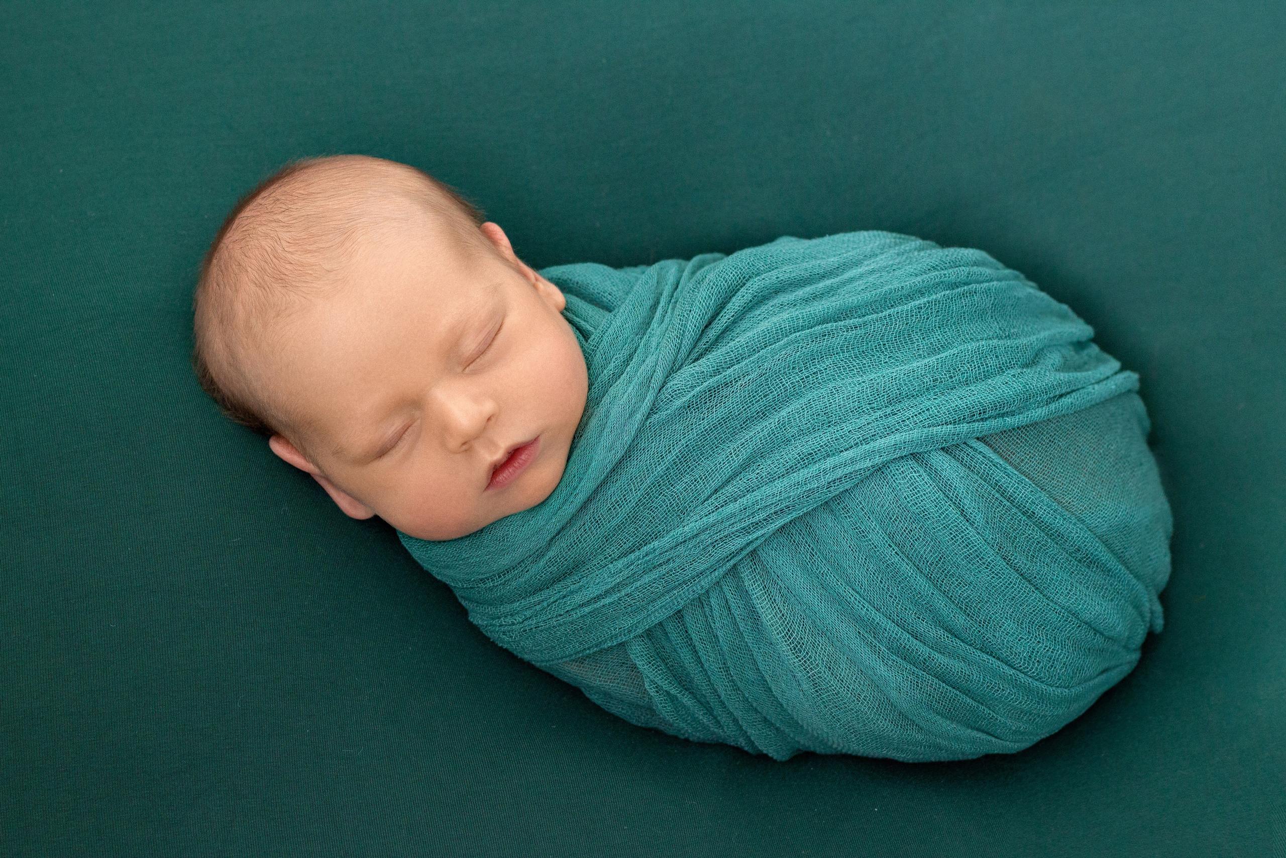 укутанный в зеленую марлю малыш спокойно спит на спинке на изумрудном фоне