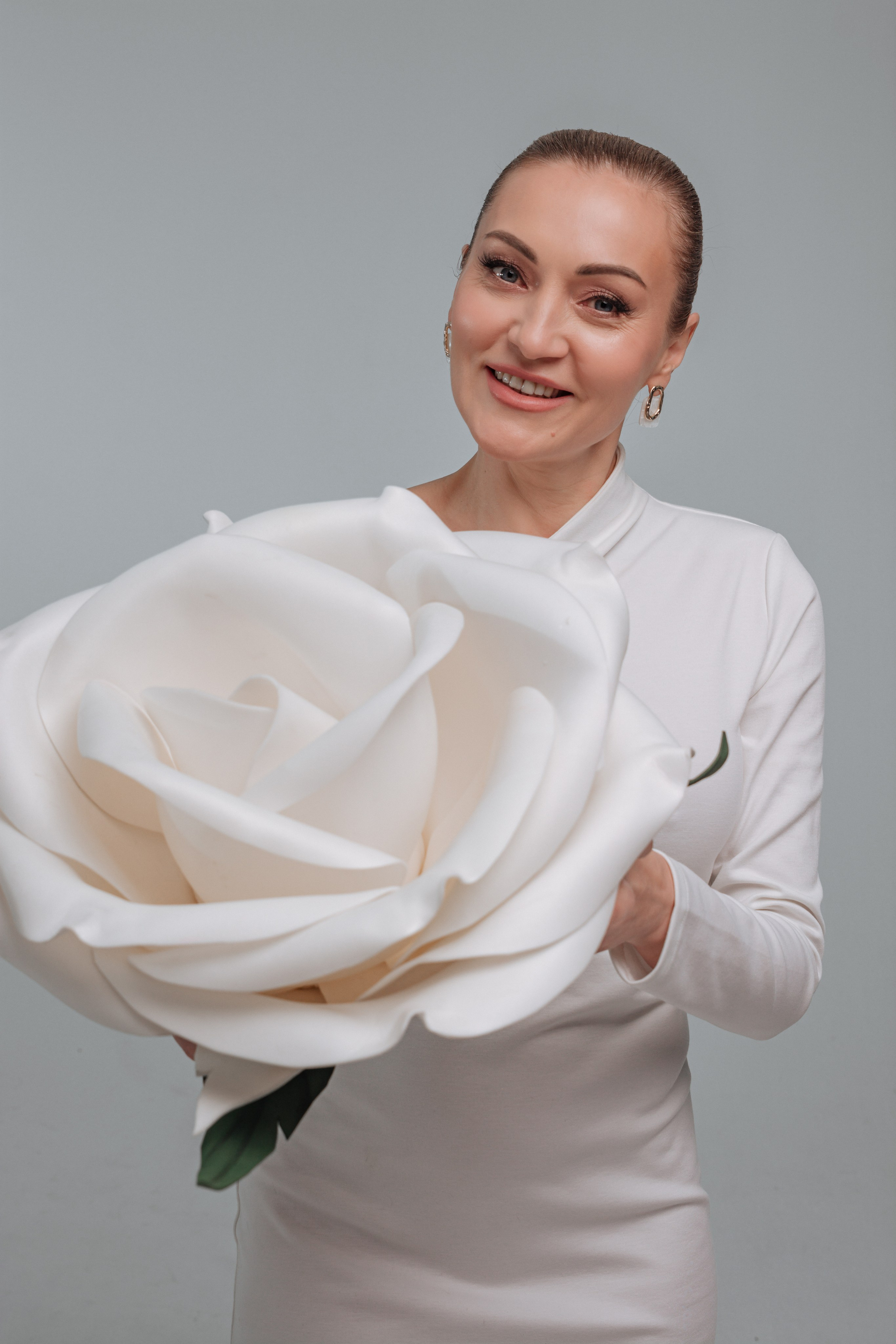 Ее лицо озарено мягкой улыбкой, взгляд устремлен вдаль, словно она погружена в приятные мысли. Окружающий ее фон размыт, акцентируя внимание на центральной фигуре.