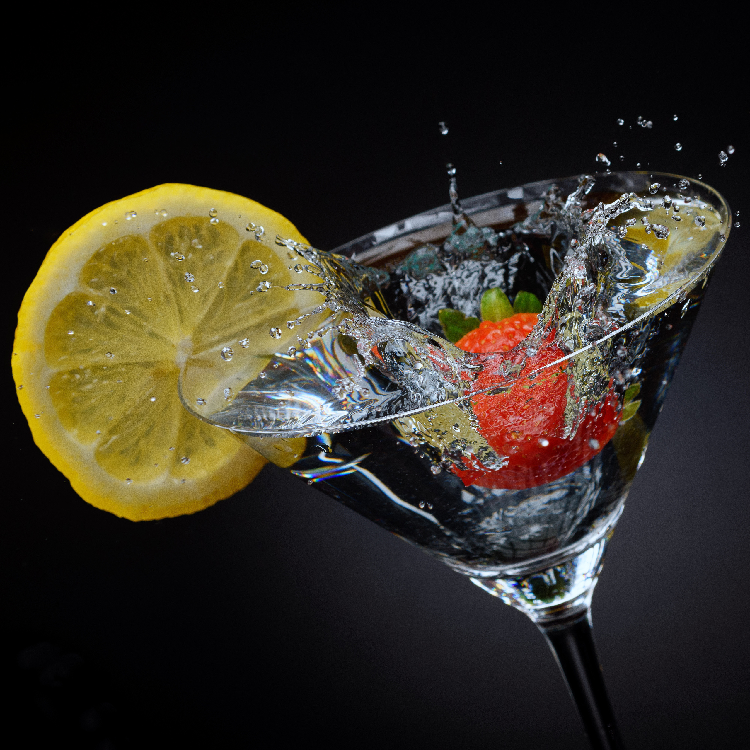бокал мартини с лимоном и падающей клубникой в воду на черном фоне