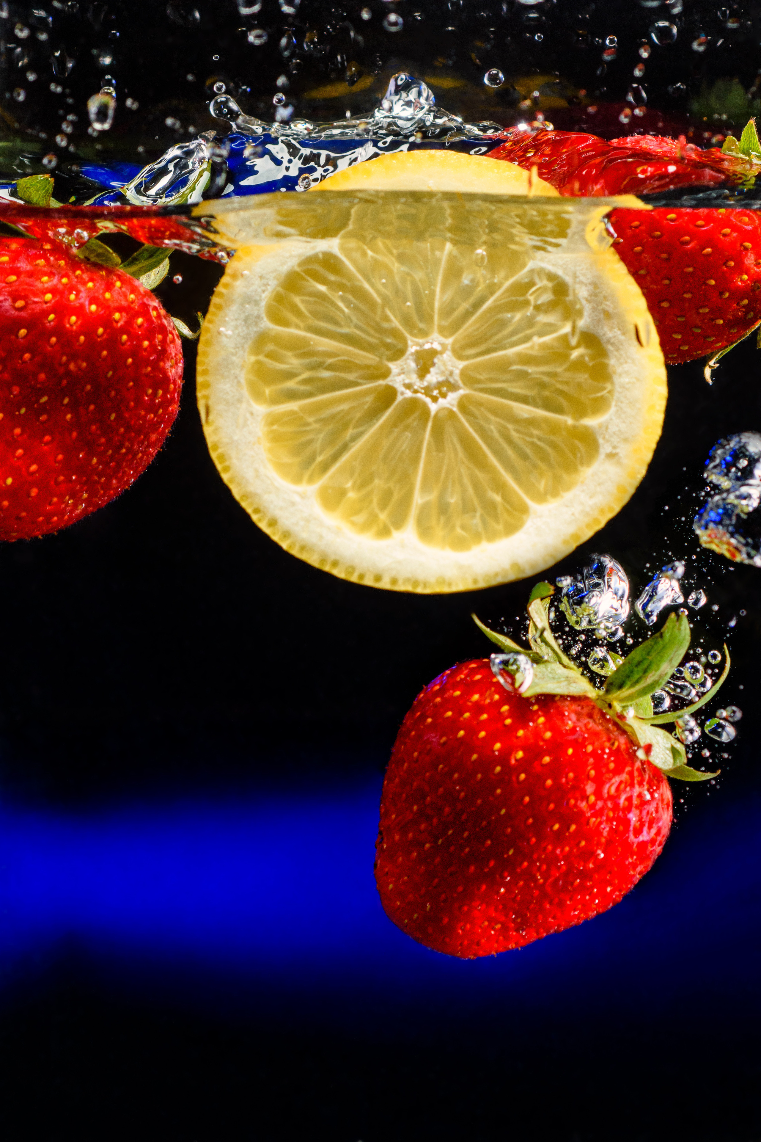 клубника, лимон падают в воду на черном фоне с синим светом