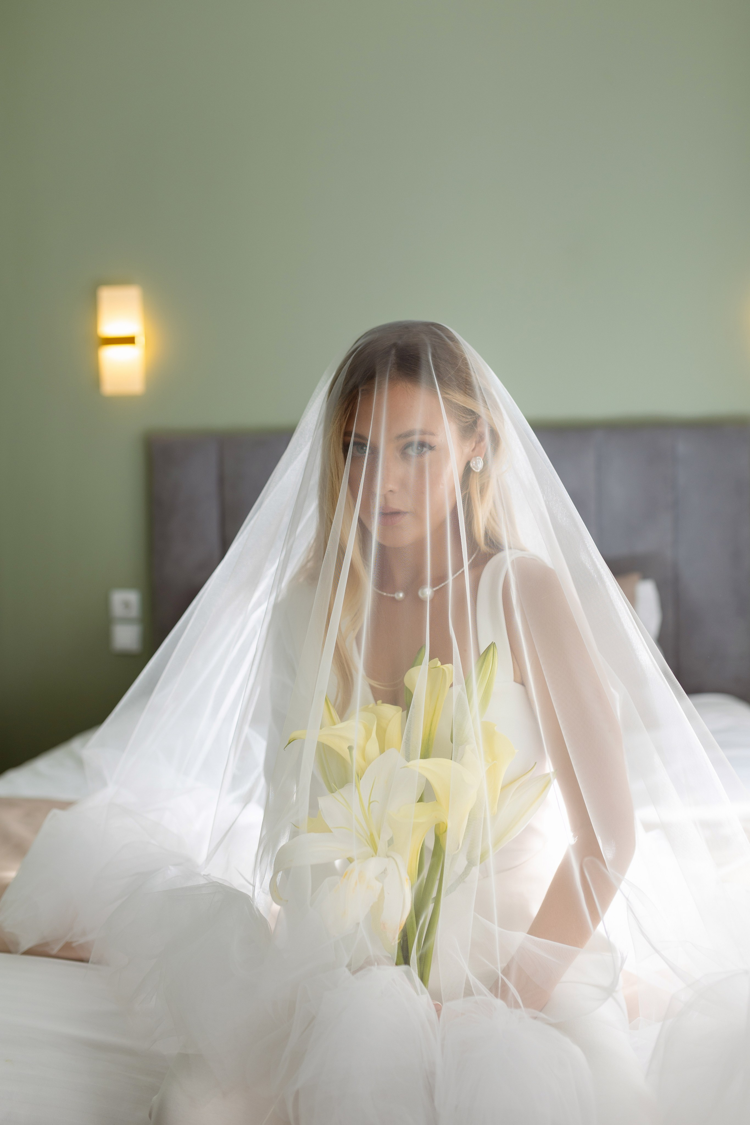 Невеста сидит на кровати покрытая белой вуалью.