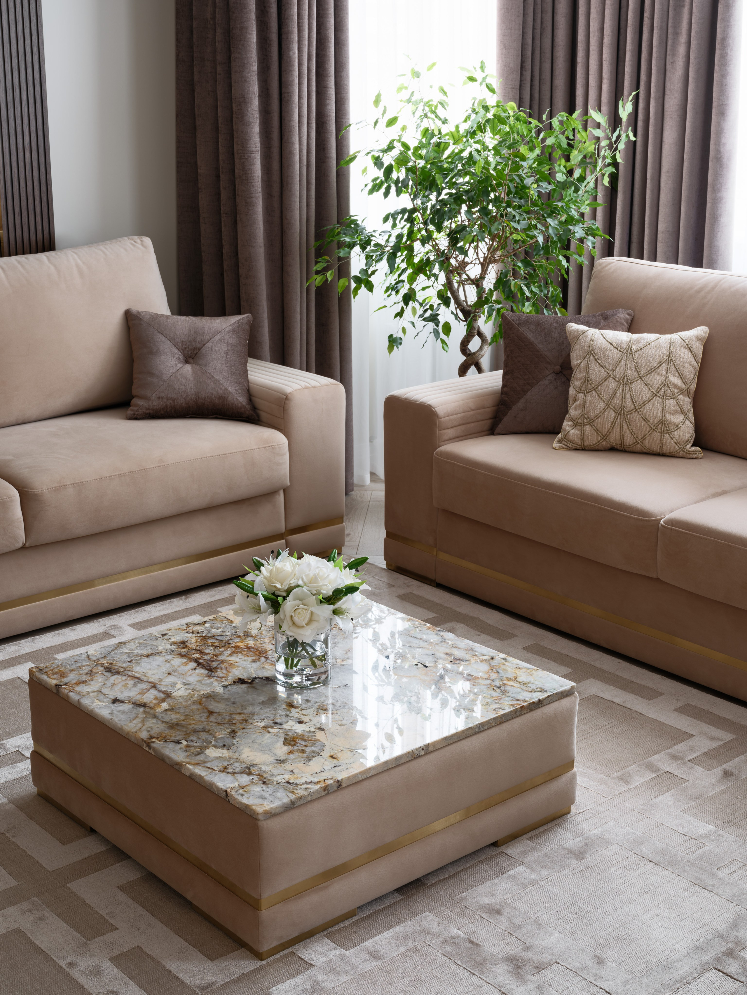 Интерьер гостиной с двумя бежевыми диванами, квадратным журнальным столиком с каменной столешницей и золотистыми деталями, ковром с геометрическим узором и живым растением у окна, создающим уютную атмосферу.