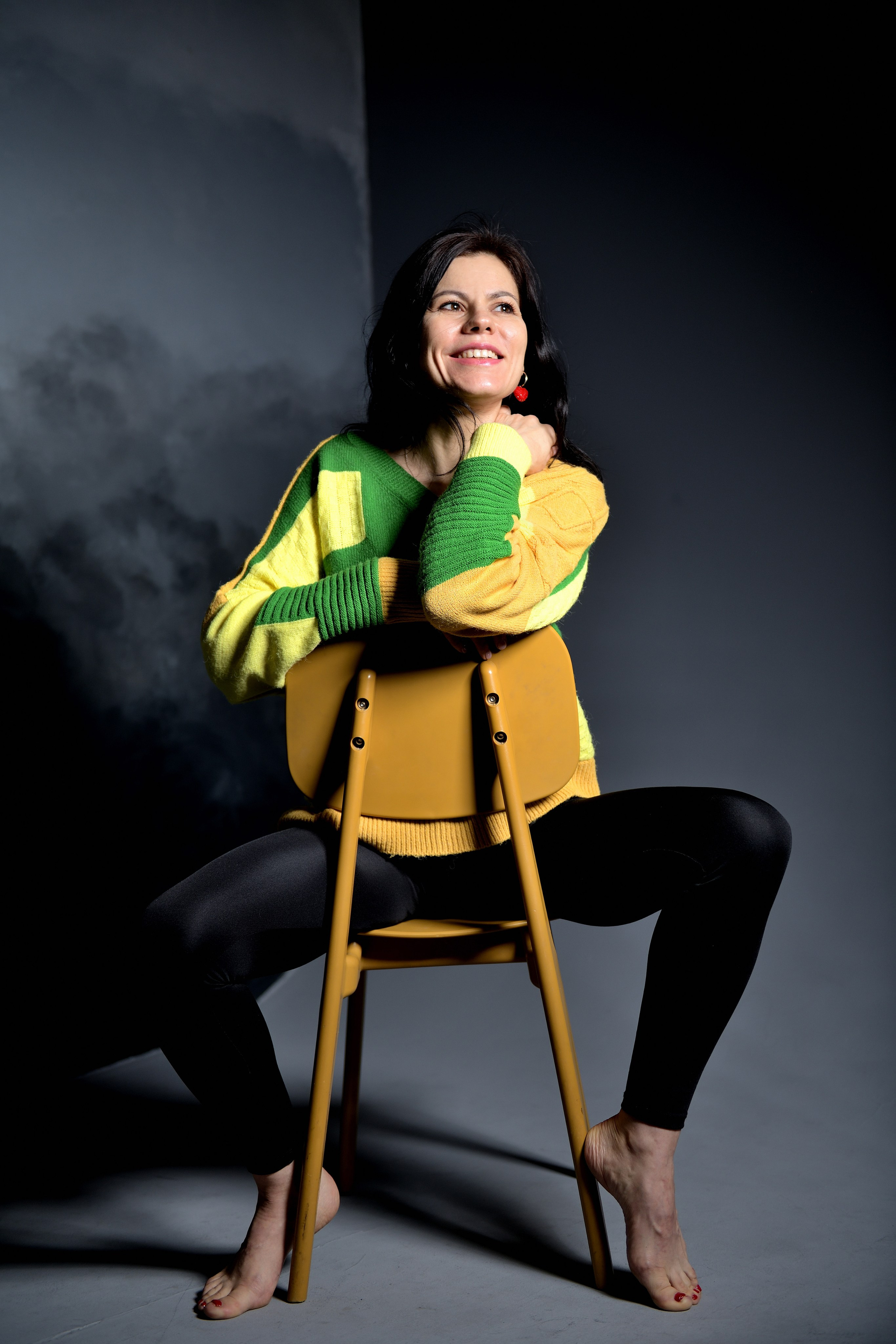 Девушка в желто-зеленом свитере сидит на деревянном суде на фоне серой стены в студии.