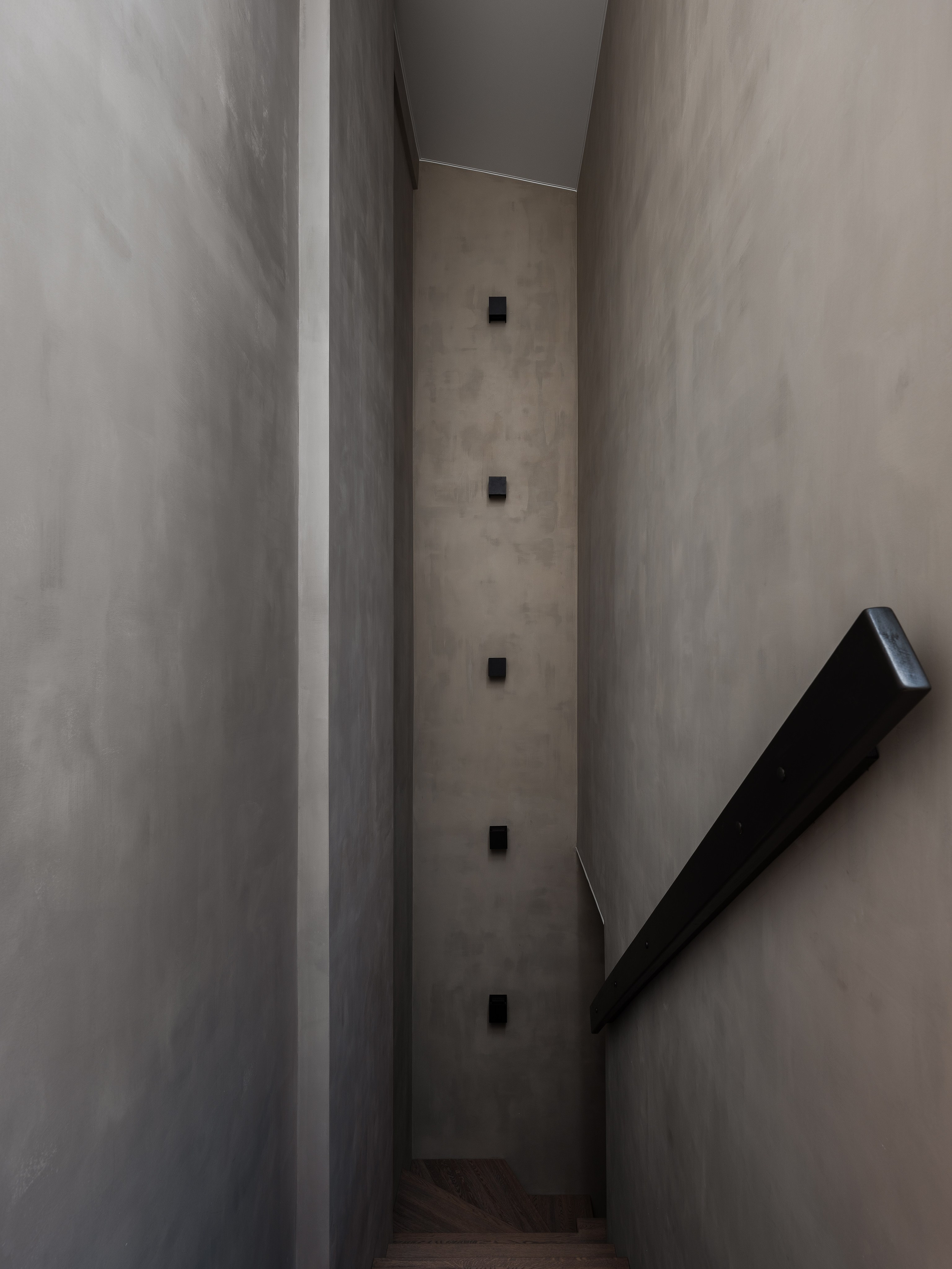 Строгий интерьер лестничного пролёта с серыми стенами, чёрными квадратными бра и деревянными ступенями, подчёркивающими атмосферу минимализма. Работа дизайнера Екатерины Мищенко, фотограф Сергей Денисенко.