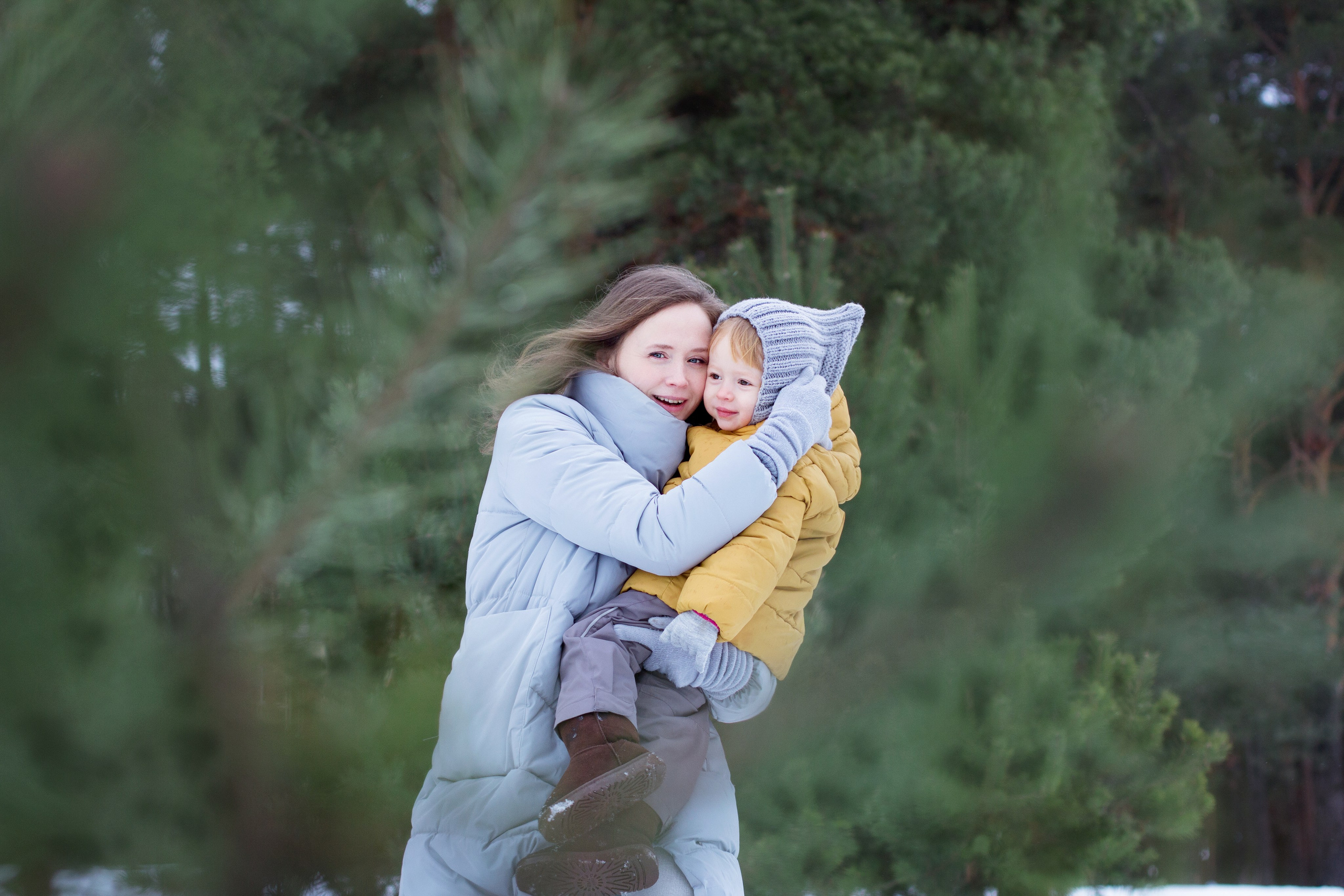 Мама держит дочку на руках в зимнем сосновом лесу