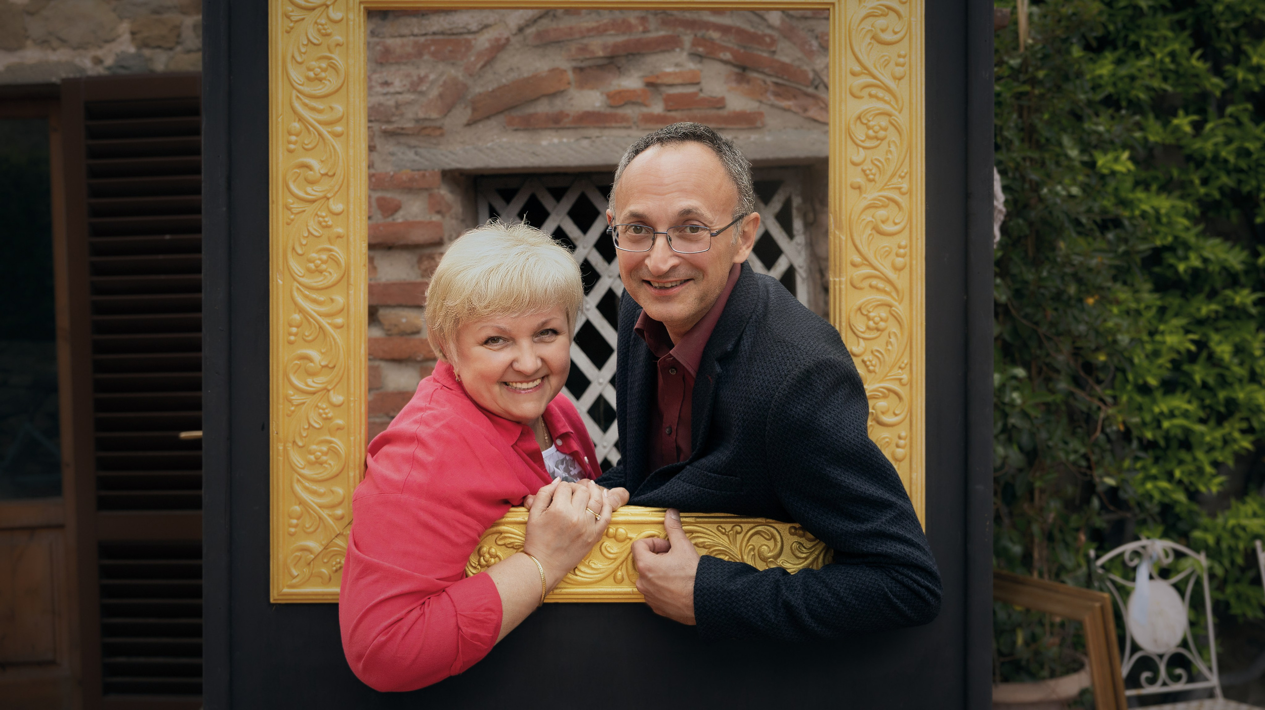 Веселый портрет пары, позирующей внутри большой, богато украшенной золотой рамы для картины, на фоне грубой кирпичной стены и зеленого плюща.