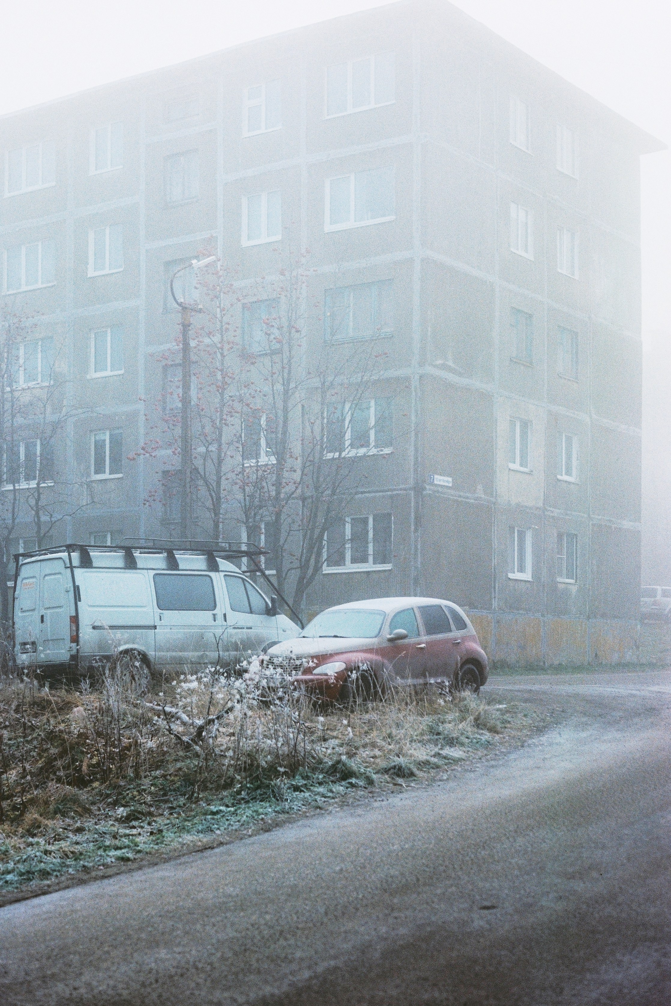 Мрачный городской пейзаж, туманный вид на пятиэтажную панельку, перед которой стоят заиндевевшие автомобили. Кировск, Россия