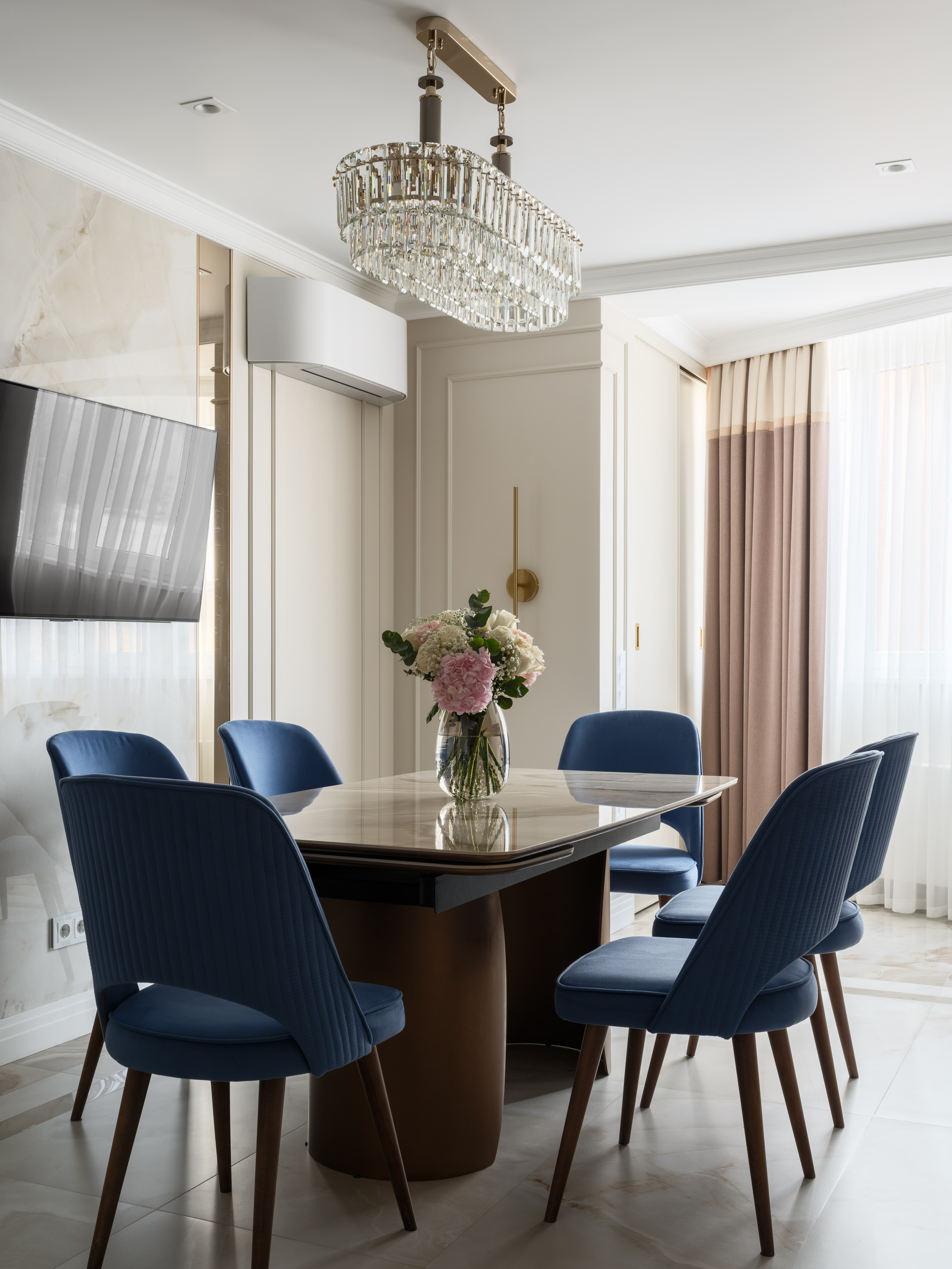 Интерьер кухни-столовой с обеденной зоной, где сочетание классического стиля и современного минимализма подчеркнуто синими стульями, массивной хрустальной люстрой, акцентными шторами и современным кондиционером, работа дизайнера Евгении Ермошиной, фотограф Сергей Денисенко.