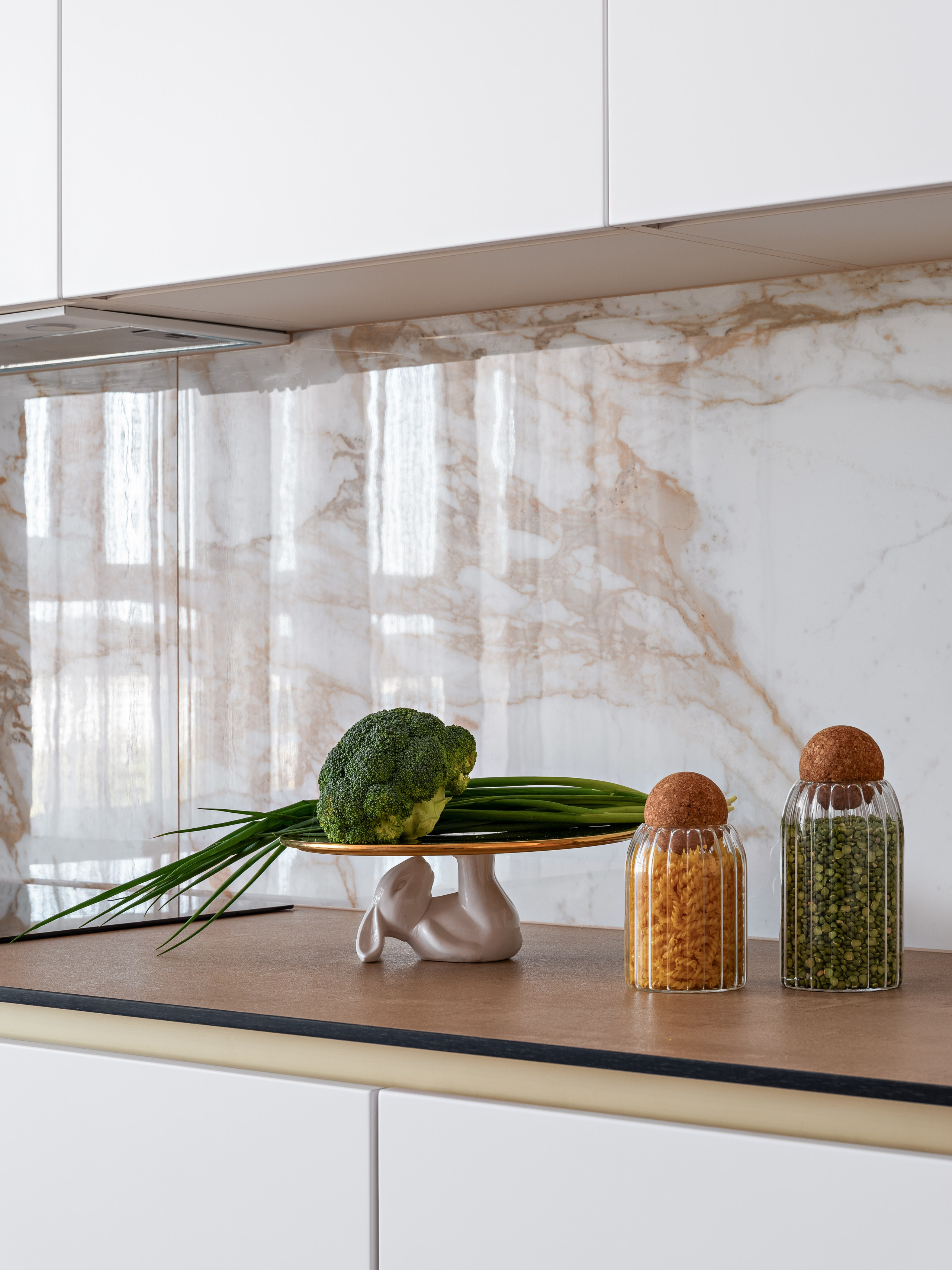 Современная кухня с белыми фасадами без ручек, столешницей под камень и мраморным фартуком, стильные баночки для хранения круп и овощи на декоративной подставке; интерьер разработан дизайнером Натальей Бунеевой, стилист Наталья Якушева, фотограф Сергей Денисенко.