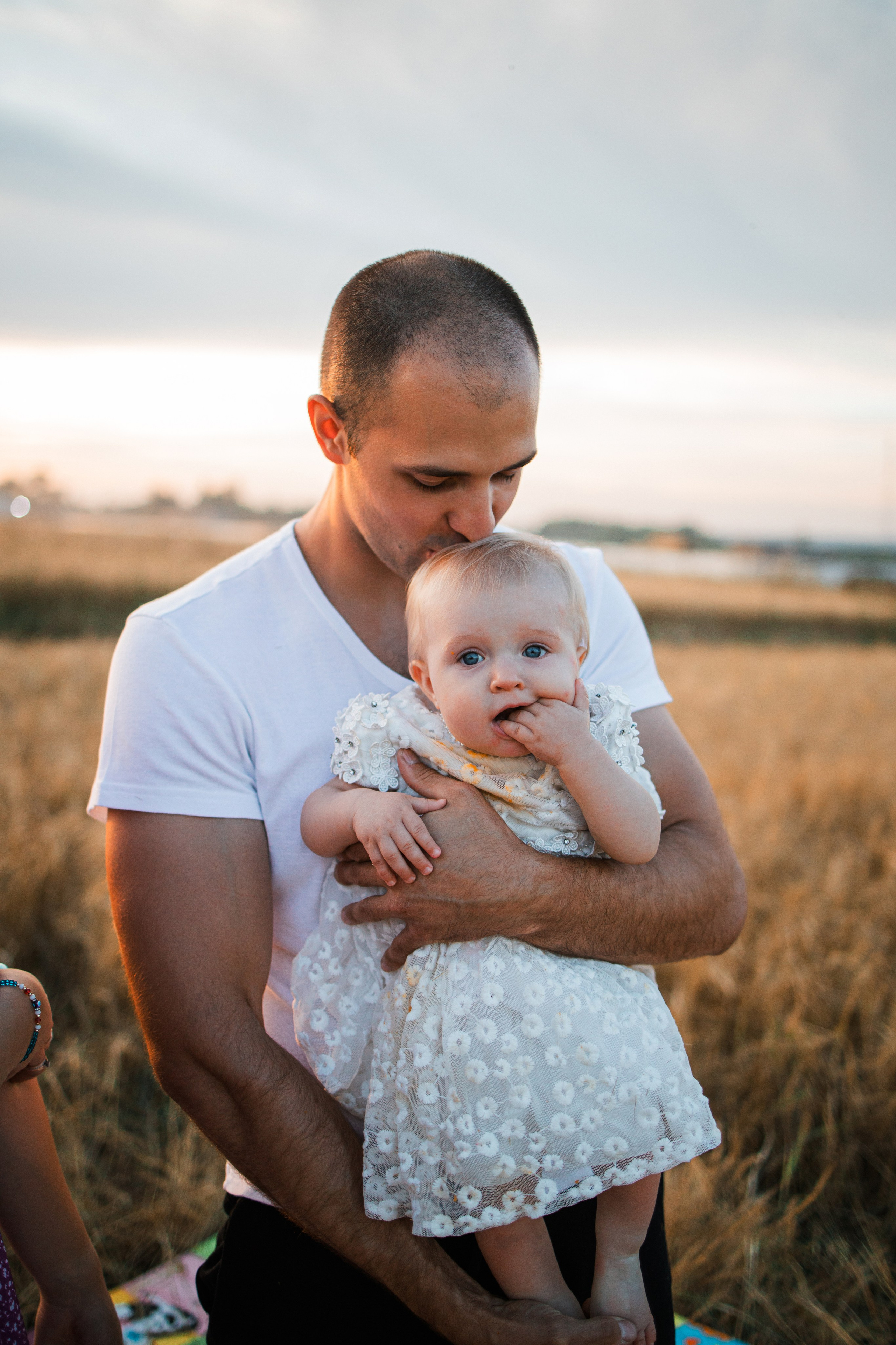 Папа целует дочку, мама улыбается рядом на пикнике в поле при закате.
