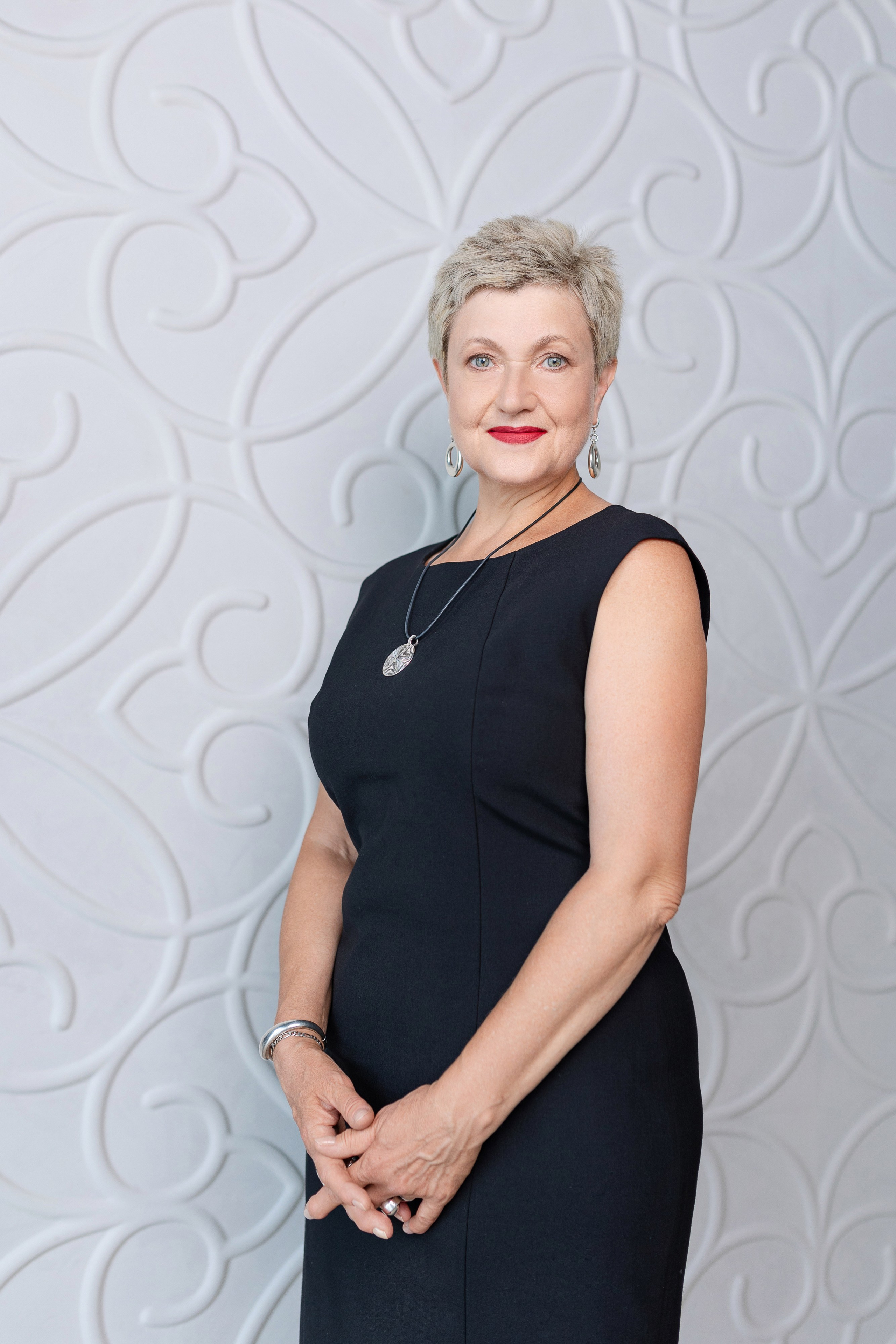 Женщина в черном платье стоит на фоне белой стены.