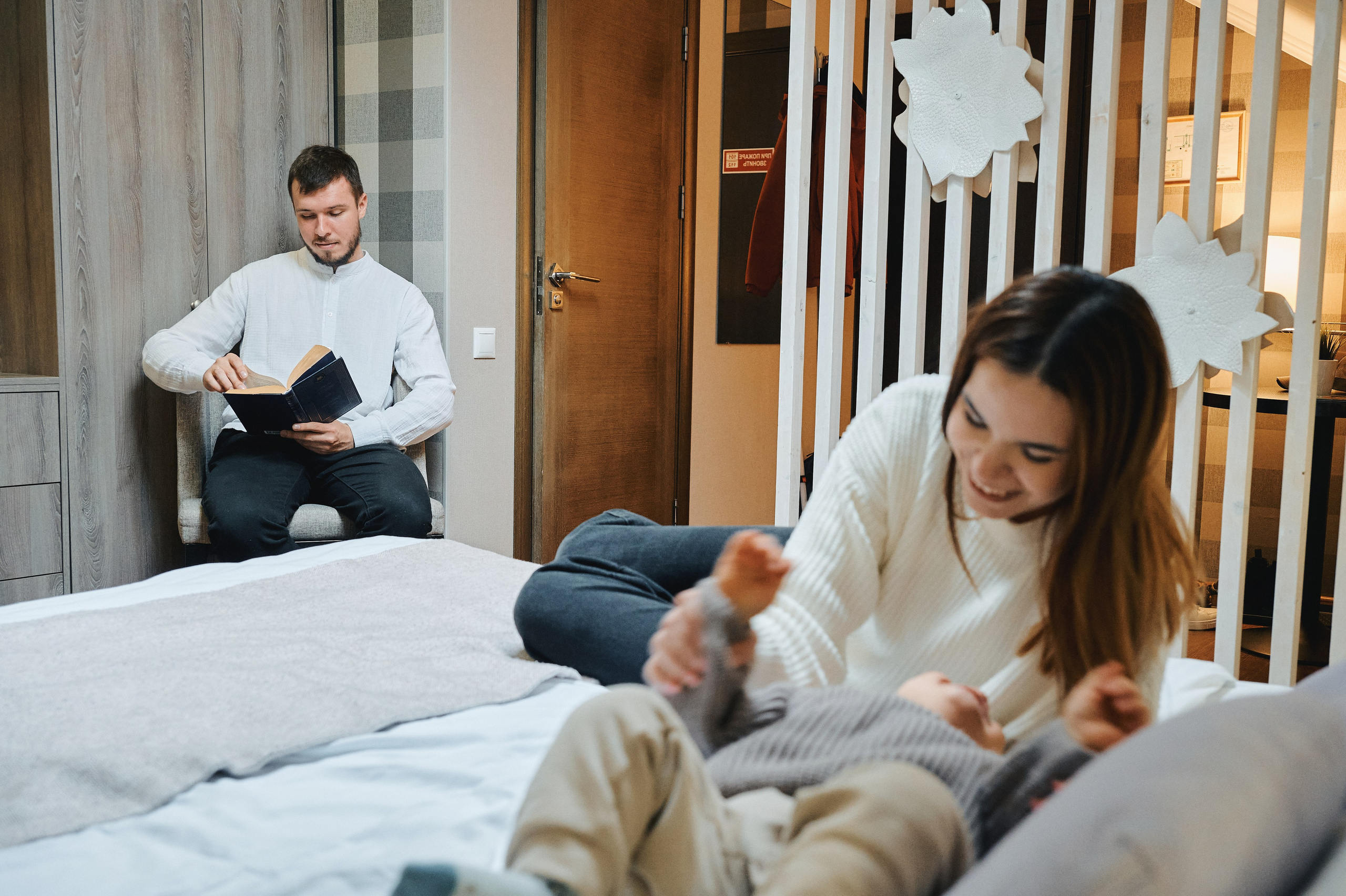 Отец читает книгу, мать играет с ребёнком на кровати в уютном номере.