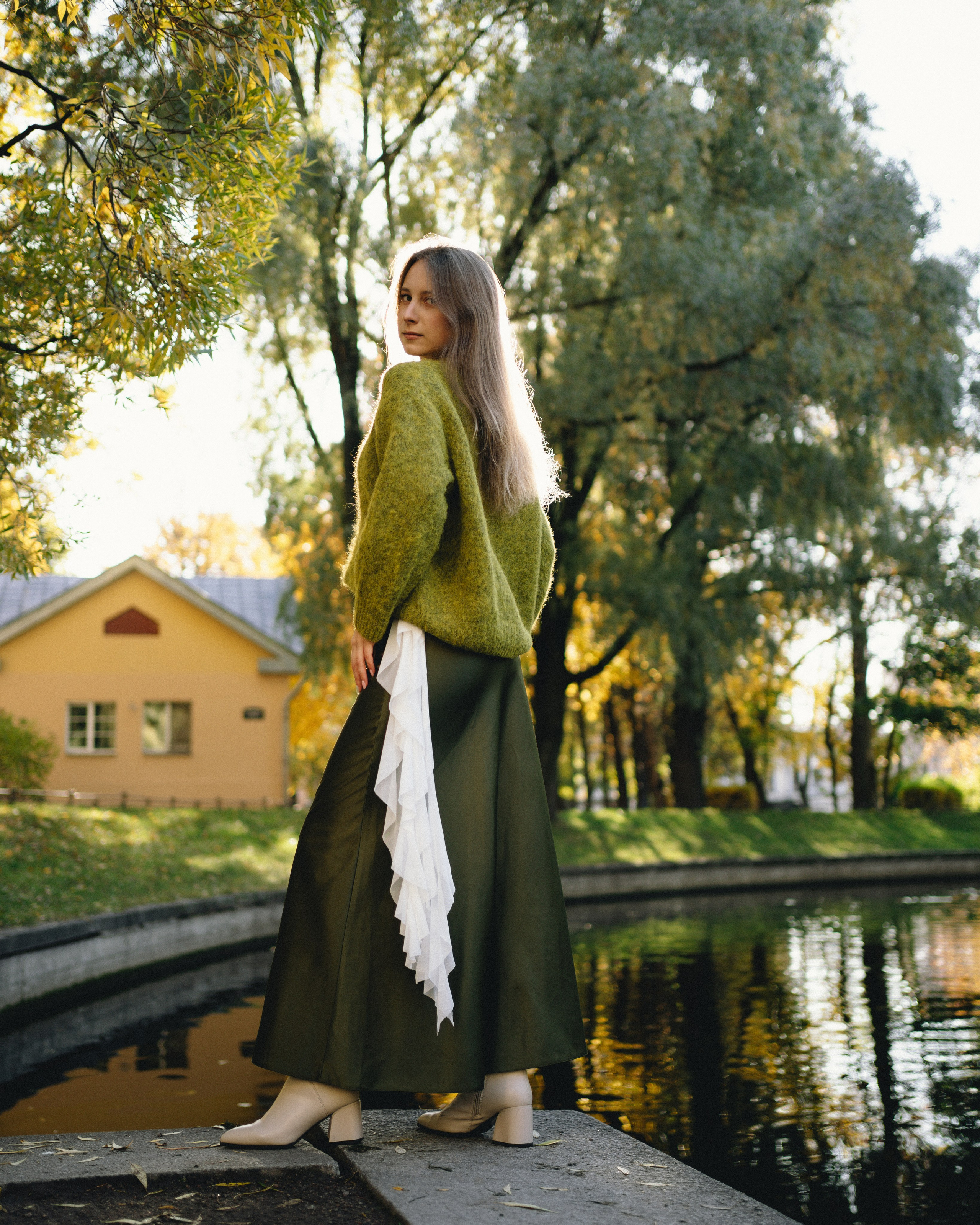 Фотография, на которой Девушка в зеленой юбке и зеленой кофте позирует в парке, на фоне пруд и осенние деревья