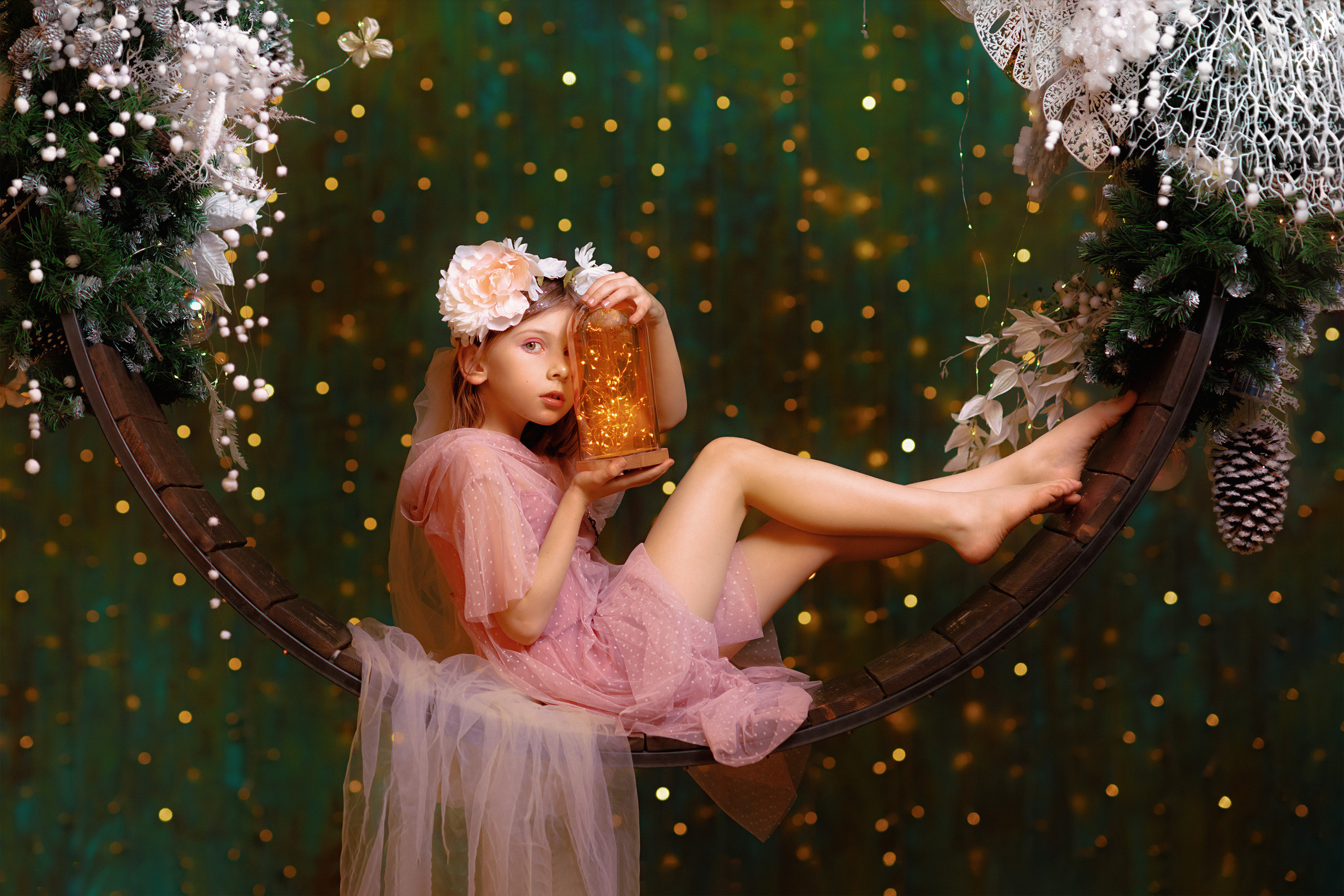 Девочка в сказочном образе с венком на голове и банкой светлячков в руках на фоне огоньков качается на волшебных качелях